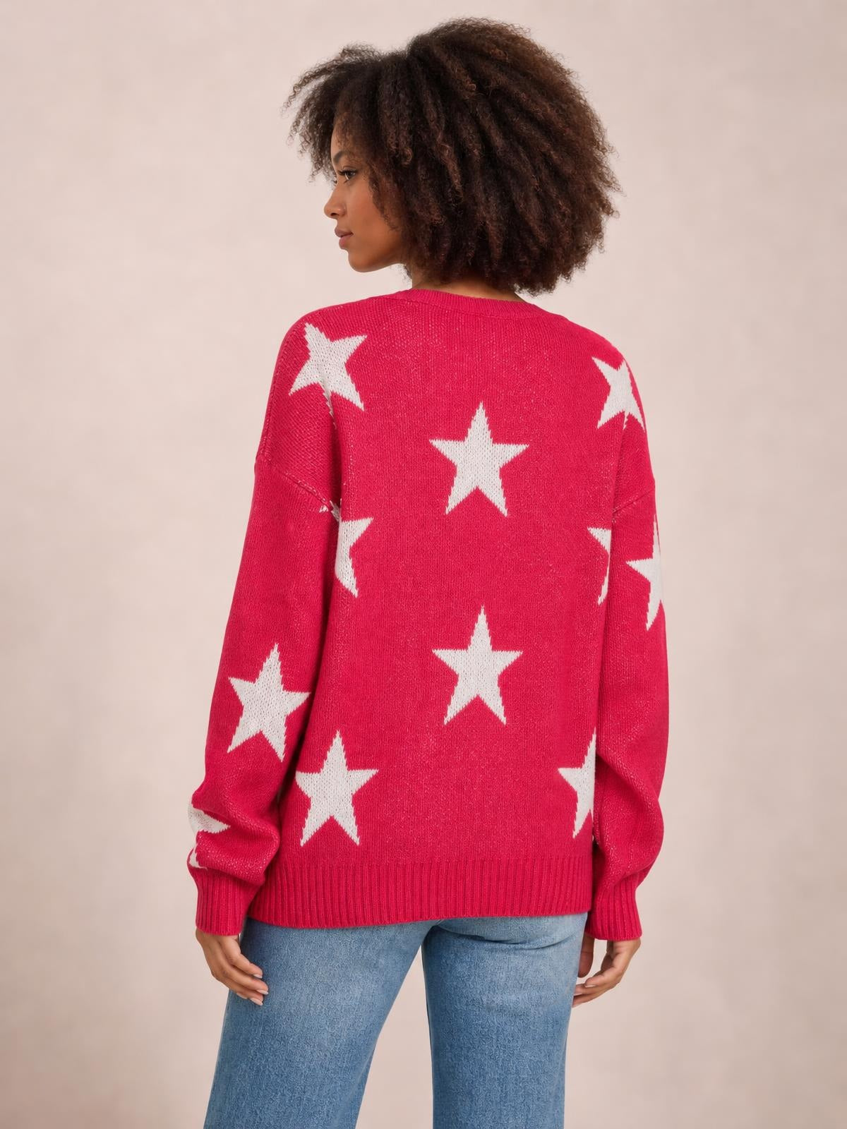 A model stands facing away in a bright pink star sweater with a round neck, dropped shoulders, relaxed silhouette, long sleeves, and ribbed cuffs and hem.