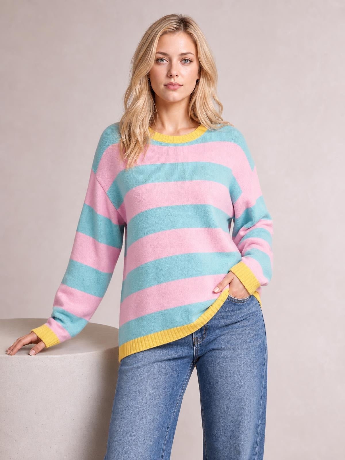 Model poses in a pastel pink and turquoise striped round-neck sweater with dropped shoulders, ribbed yellow trim, long sleeves, and a relaxed silhouette.
