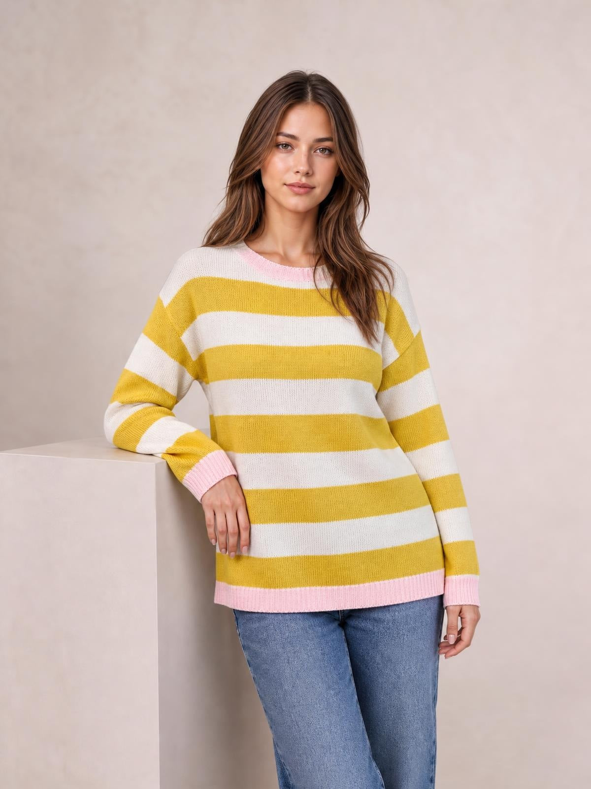 Model wearing a relaxed striped round neck dropped shoulder sweater in mustard yellow and white with pink ribbed cuffs and hem, leaning against a pedestal.