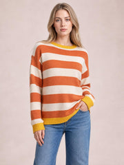 Model wearing an orange and ivory striped round-neck sweater with dropped shoulders, a relaxed silhouette, and contrasting yellow ribbed cuffs and hem.