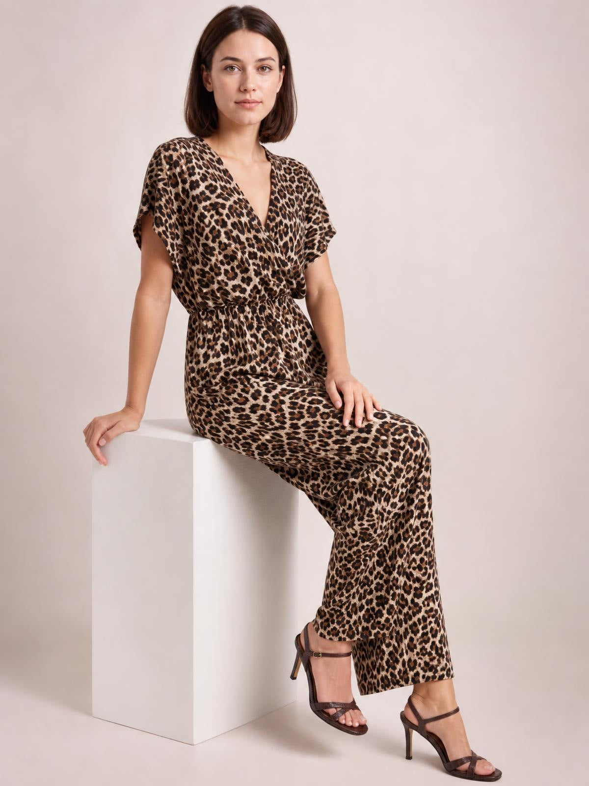 Model seated in a leopard-print wide-leg jumpsuit with a V-neck, short sleeves, elasticized waist, and relaxed flowing silhouette.