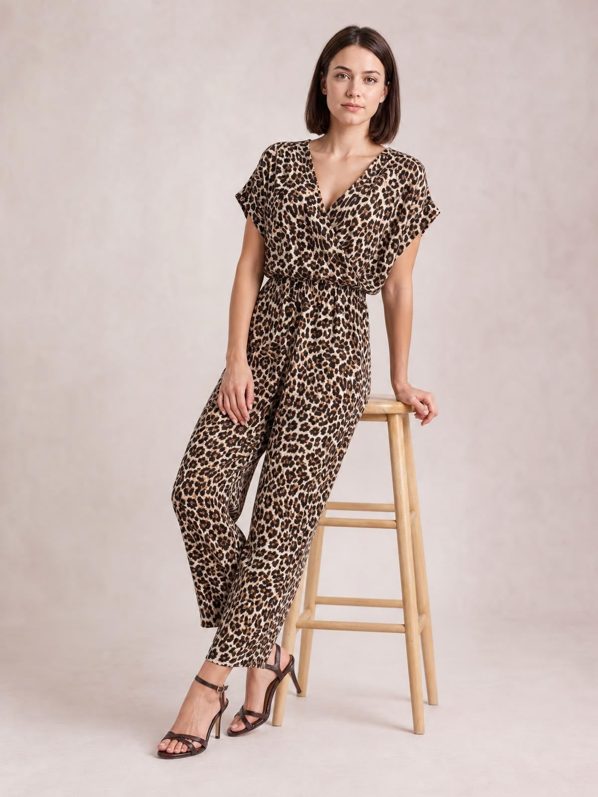 Model seated on a stool wears a brown and black animal print wide leg jumpsuit with a V-neck, short sleeves, and a defined elastic waist.