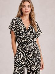 Model standing with hands in pockets wears a black-and-cream animal print jumpsuit with a V-neck, short sleeves, elastic waist, and wide-leg silhouette.
