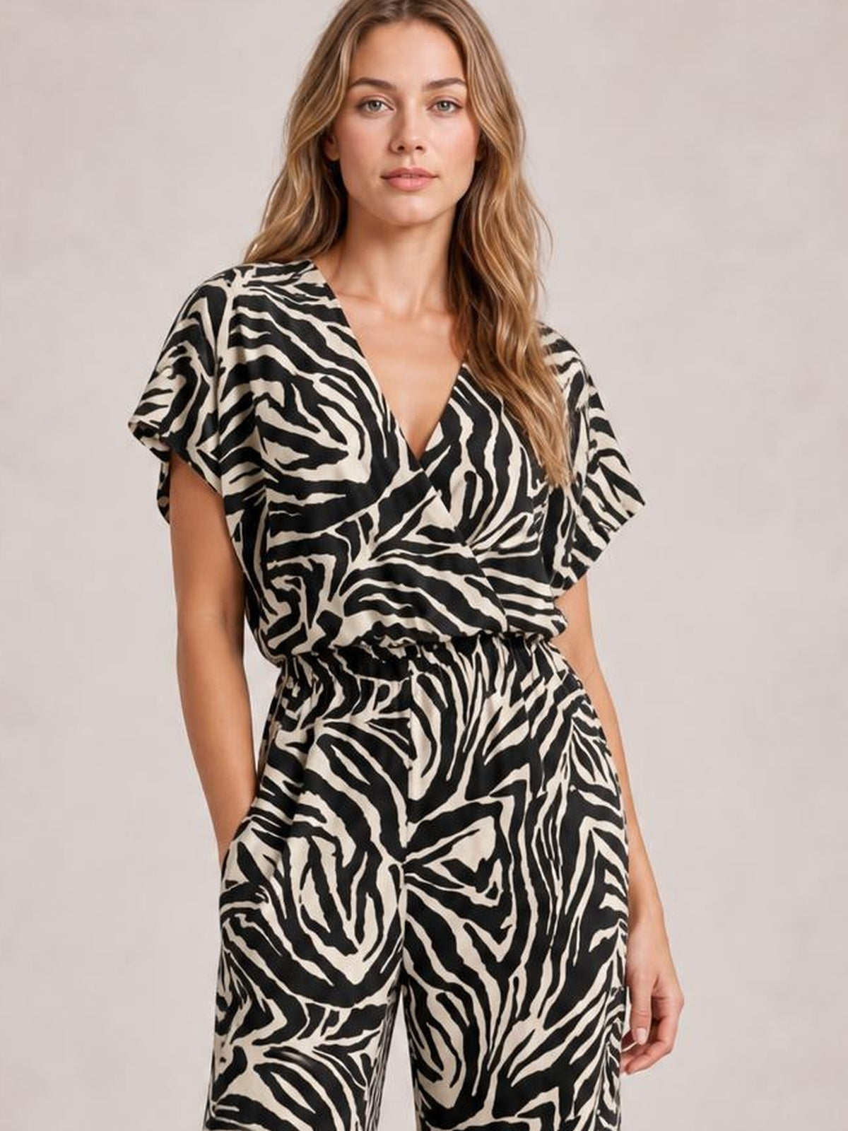Model standing with hands in pockets wears a black-and-cream animal print jumpsuit with a V-neck, short sleeves, elastic waist, and wide-leg silhouette.