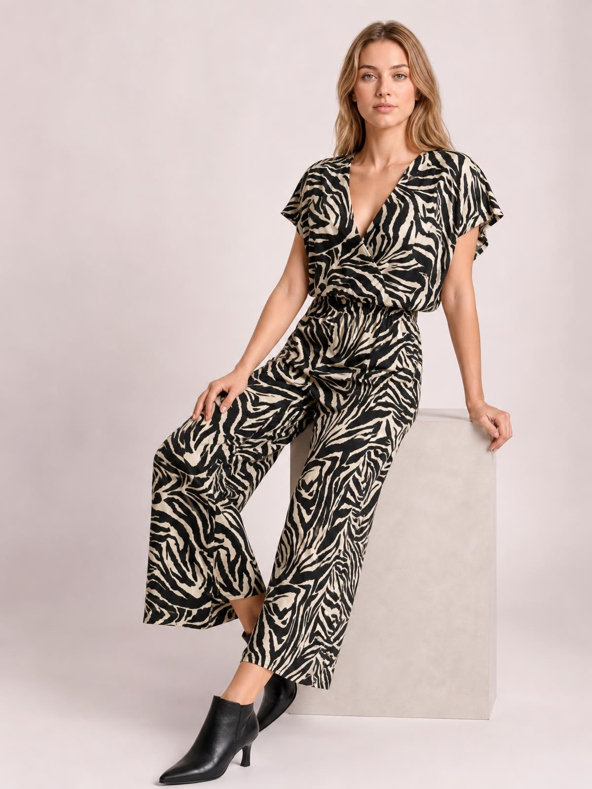 Model sits on a cube wearing a black-and-cream animal print jumpsuit with short sleeves, V-neckline, elastic waist, pockets, and wide legs.
