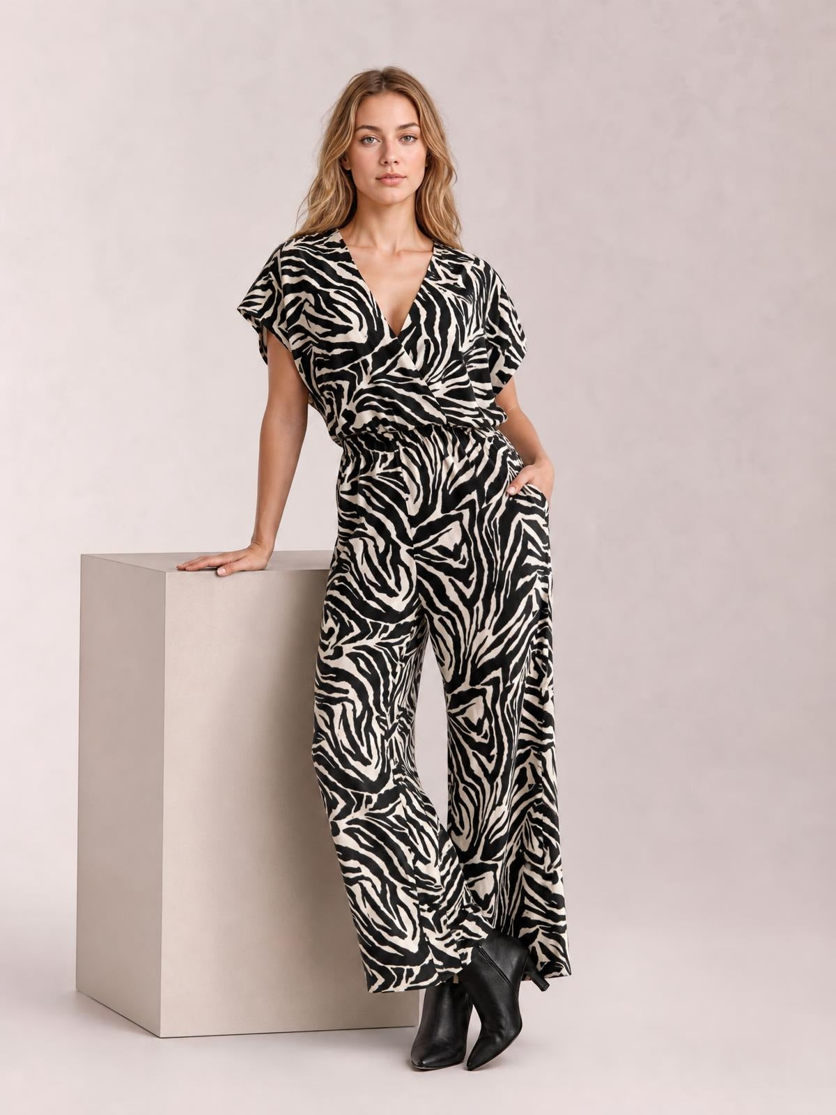 A model wears a black and cream animal print wide-leg jumpsuit with a V-neck, short sleeves, elastic waist, and pocket, leaning on a block.