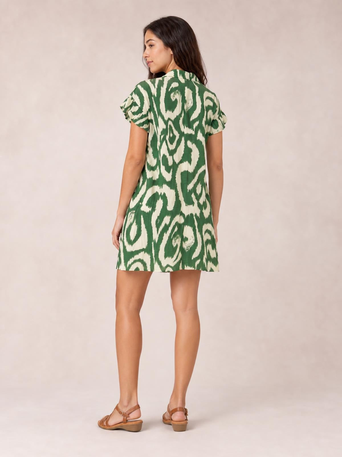 Model looks over her shoulder in a green and cream abstract printed shirt mini dress with a loose silhouette, collared neckline, and frill sleeves.