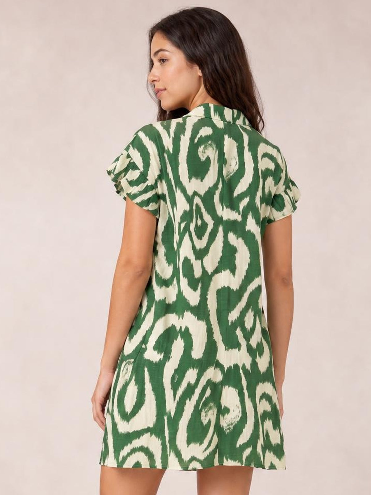 Model turned to show a green and cream abstract printed shirt mini dress with a collared neckline, frill sleeves, and a relaxed A-line silhouette.