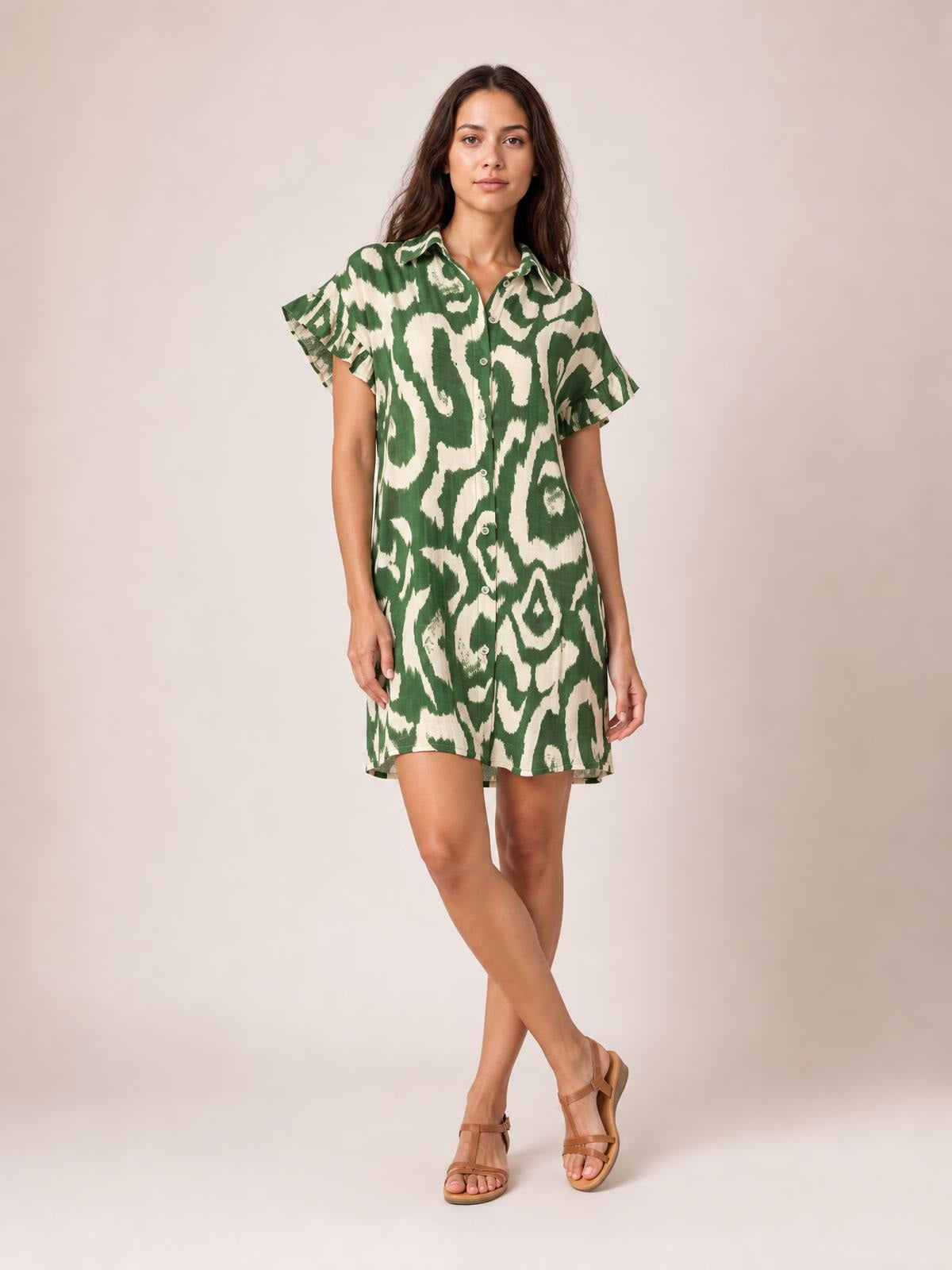 Model poses with crossed legs in a green and cream abstract printed mini dress featuring a collared button front, flared frill sleeves, and a relaxed straight silhouette.