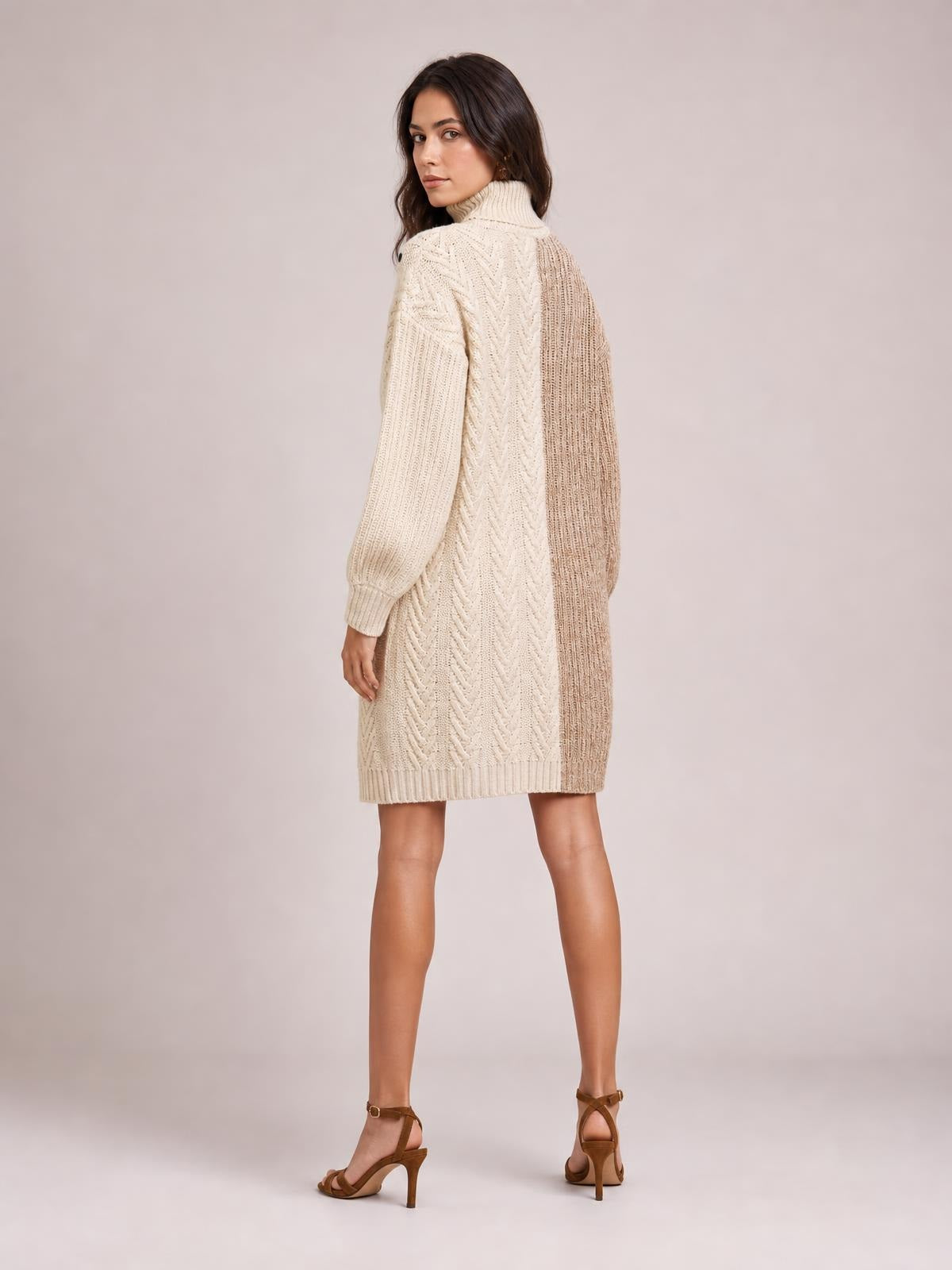 A model turns to show a cream and taupe color block turtleneck sweater dress with ribbed knit panels, long sleeves, and a relaxed above-knee silhouette.