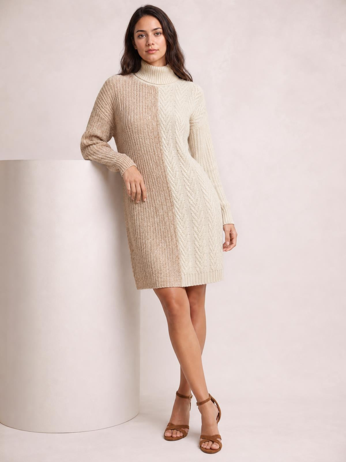 Model leans against a column wearing a beige and cream color block turtleneck sweater dress with long sleeves, ribbed knit texture, and a straight above-knee silhouette.