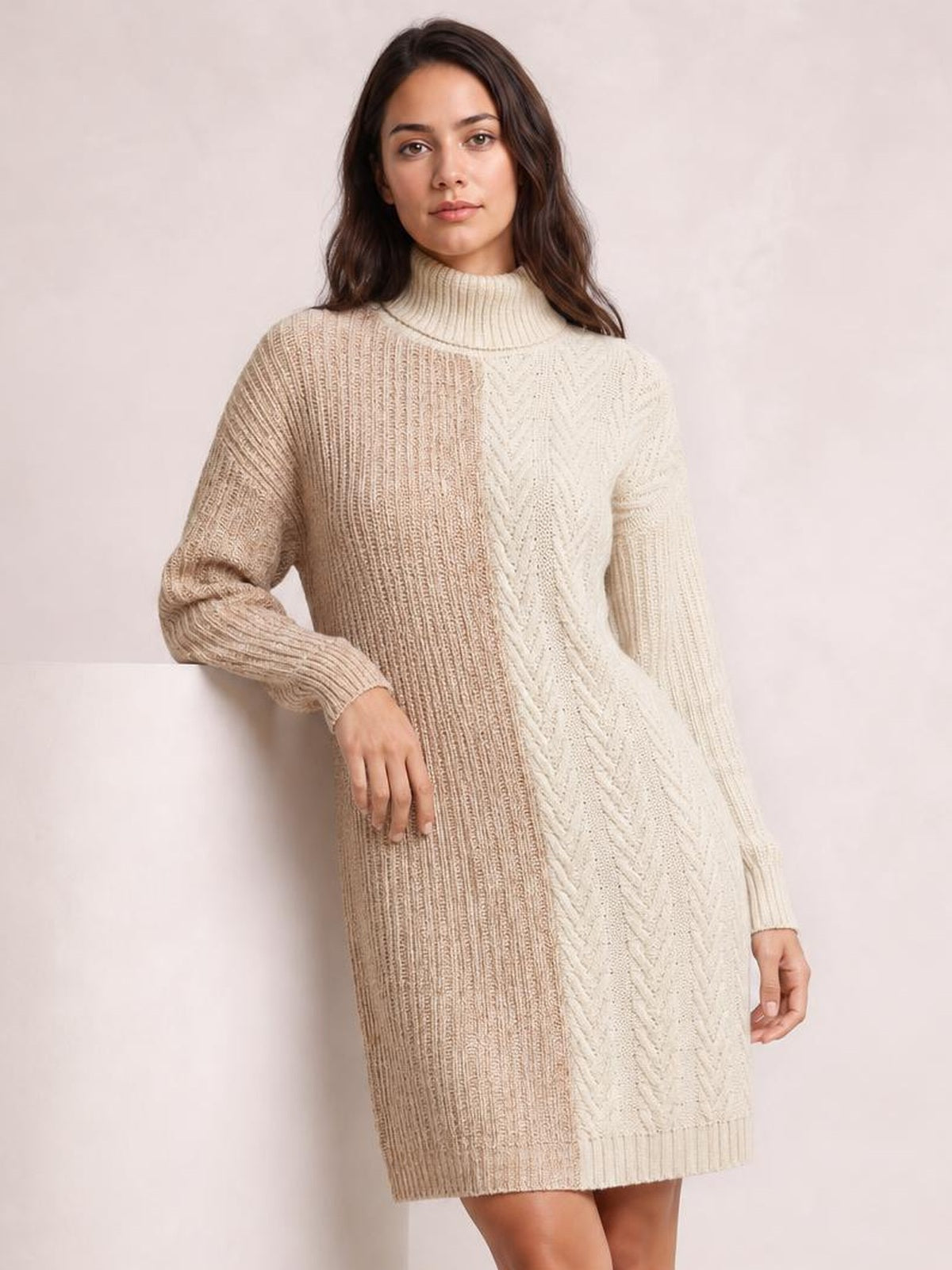 A model poses in a ribbed color block turtleneck sweater dress with tan and cream panels, long sleeves, cable-knit detailing, and a straight above-knee silhouette.