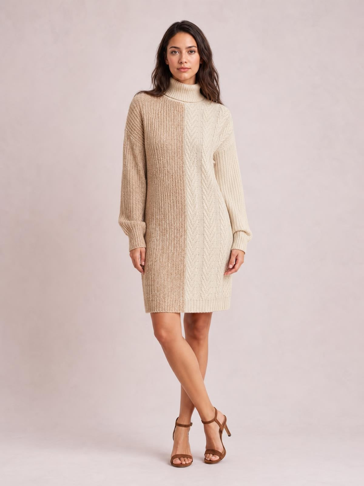 A model stands with crossed legs wearing a beige and cream color block turtleneck sweater dress with ribbed knit texture, long sleeves, and a straight silhouette.