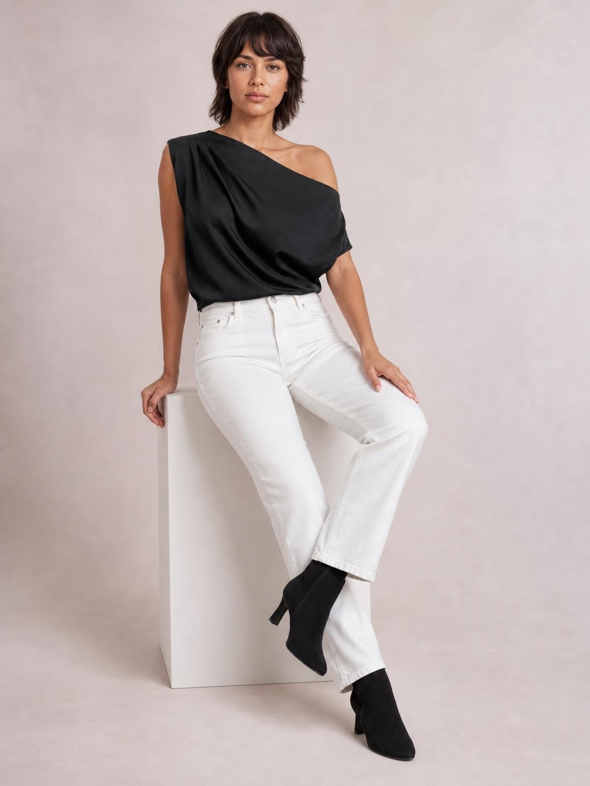 Model seated in a black asymmetrical one-shoulder draped blouse with a relaxed silhouette and gathered fabric detailing across the front.