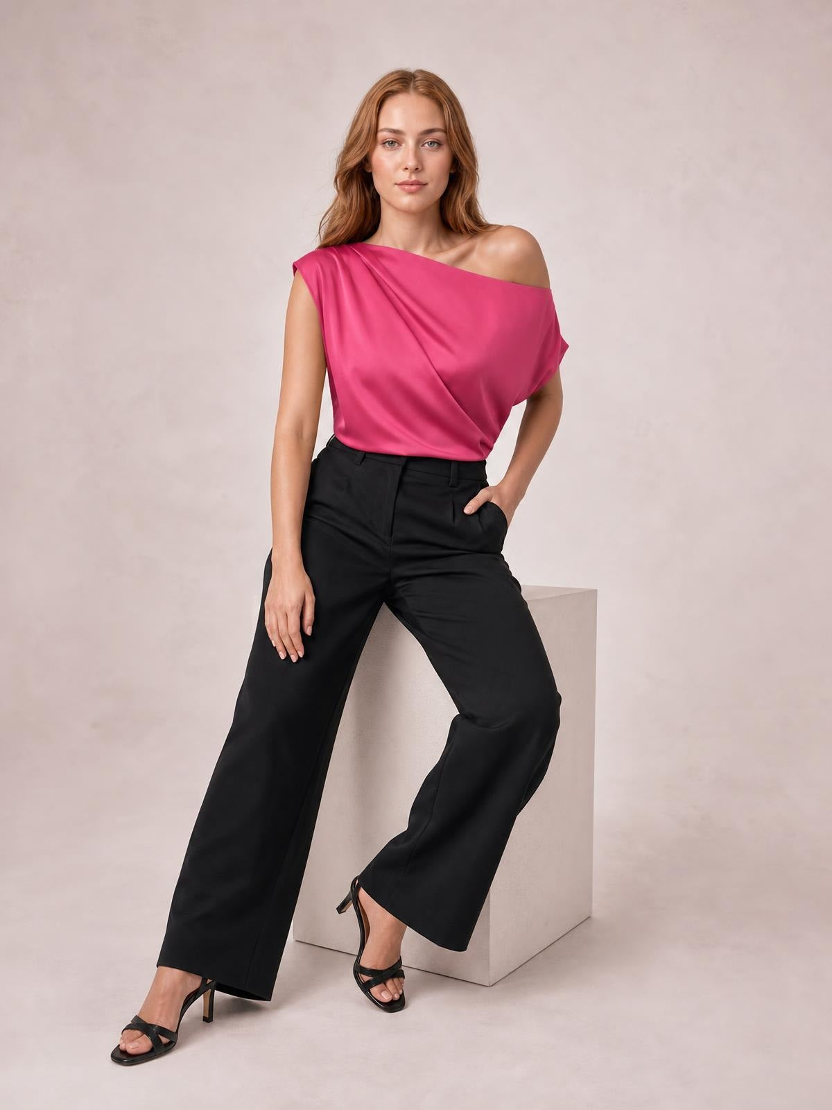 Model poses in a fuchsia asymmetrical one-shoulder draped blouse with soft folds and a relaxed silhouette, paired with black wide-leg trousers.