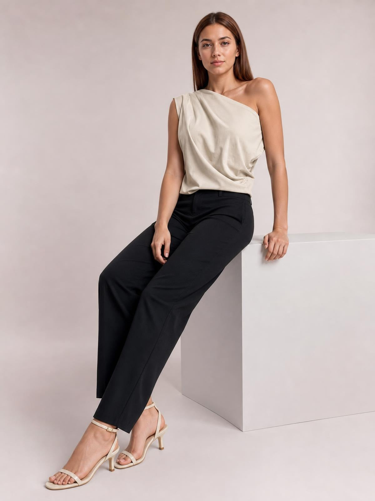 A model sits with one hand resting on a ledge, wearing a cream asymmetric one-shoulder draped blouse with soft folds and a relaxed silhouette.