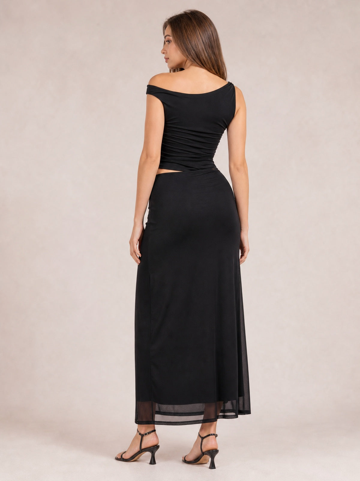 A model poses from behind in a black asymmetrical sleeveless maxi dress with twisted ruching, a waist cutout, figure-skimming silhouette, and sheer ankle-length hem.