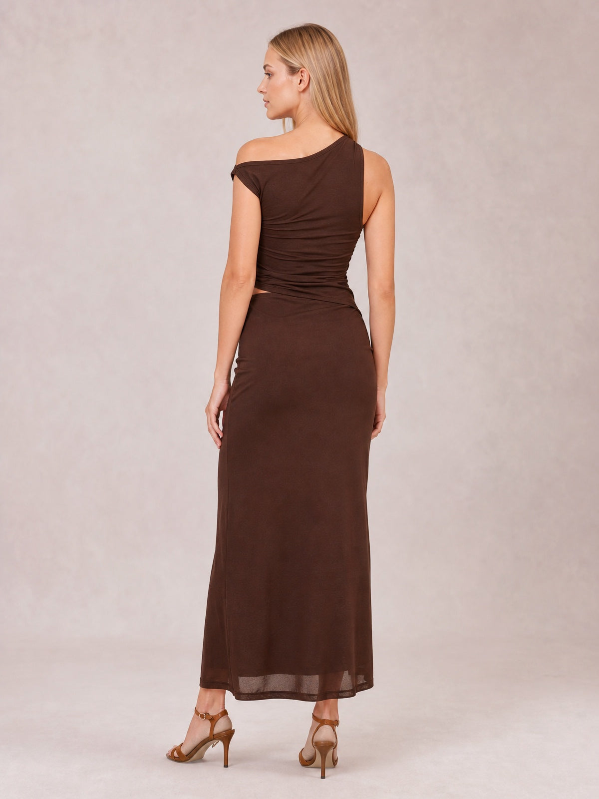 Model poses in a brown asymmetrical sleeveless maxi dress with a twisted, ruched bodice, waist cutout, figure-skimming silhouette, and ankle-length skirt.
