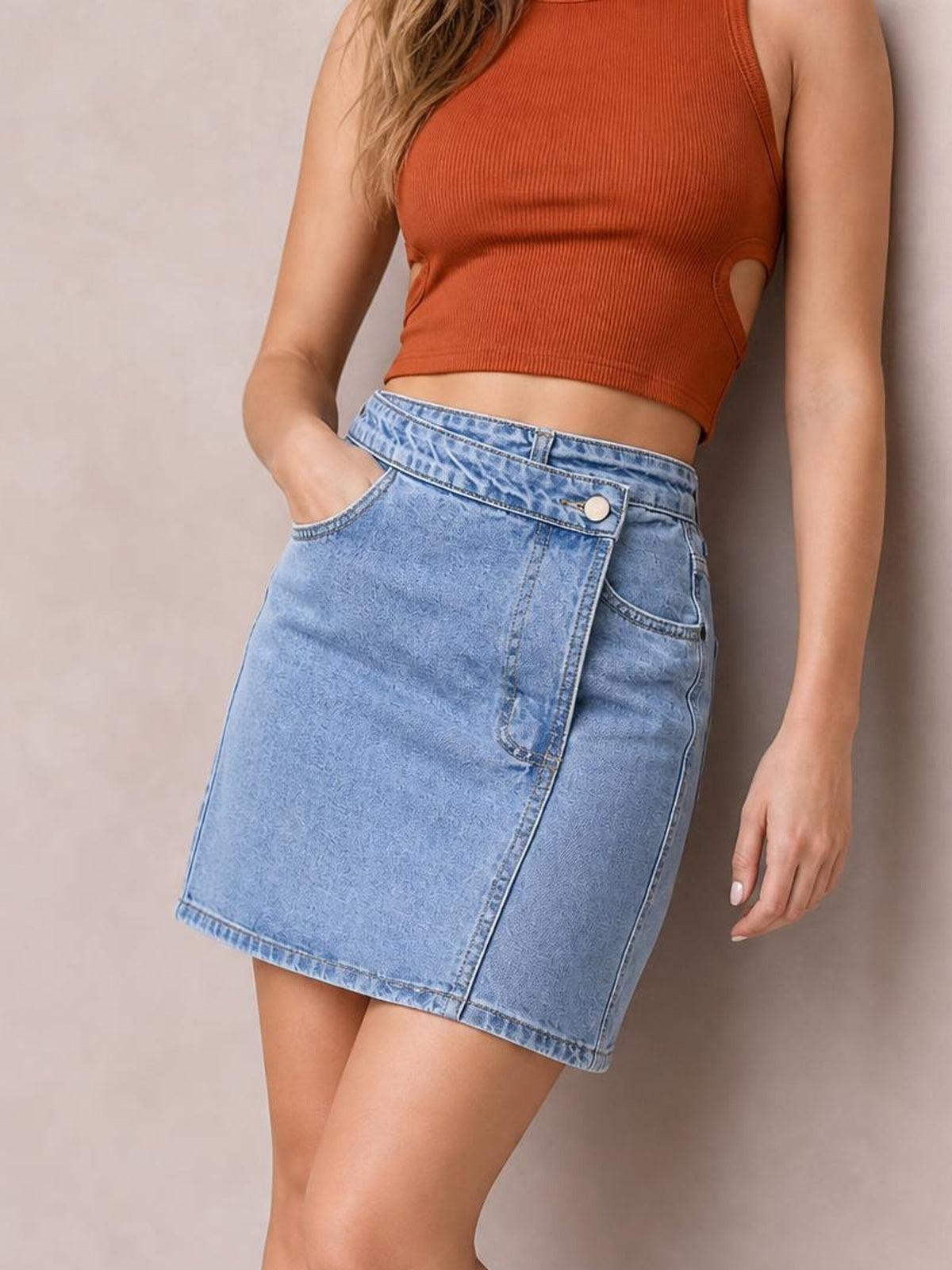 A model poses in a light blue denim mini skirt with an asymmetrical wrap front, buttoned waistband, stitched panels, and a structured silhouette.