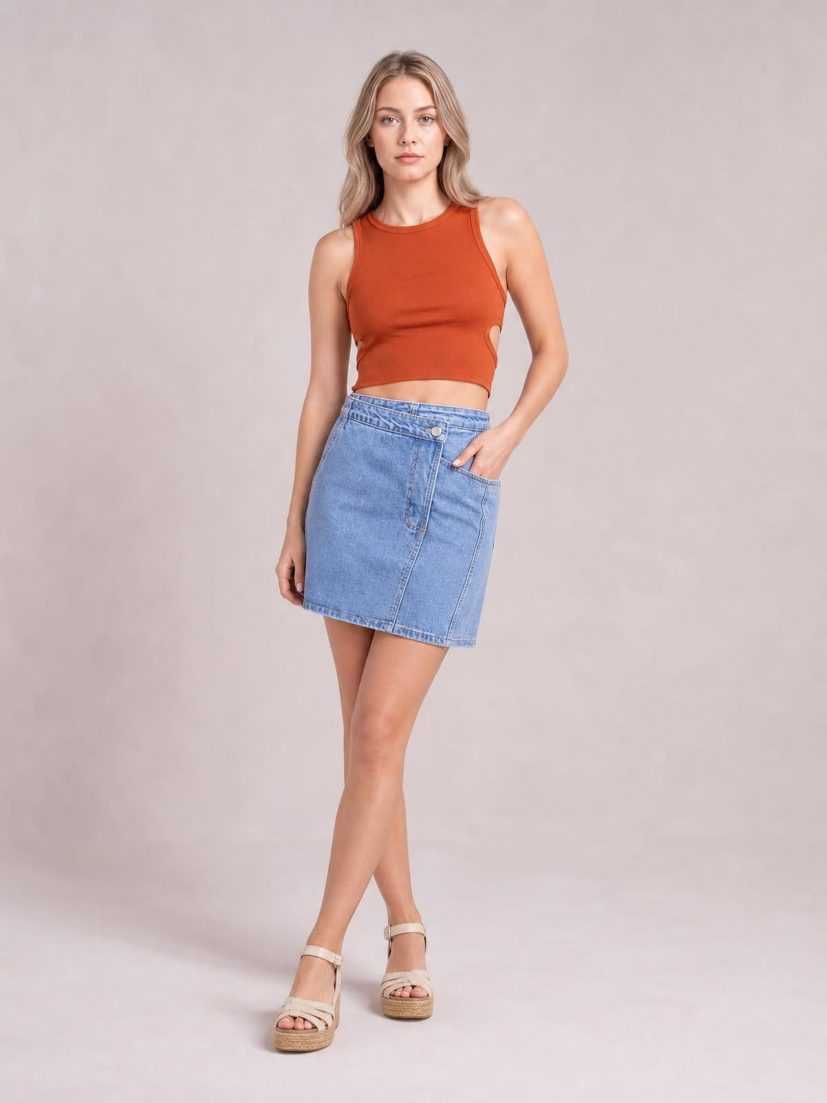 A model poses with one hand in the pocket, wearing a light-blue asymmetrical denim mini skirt with button details, a stitched front panel, and a straight silhouette.