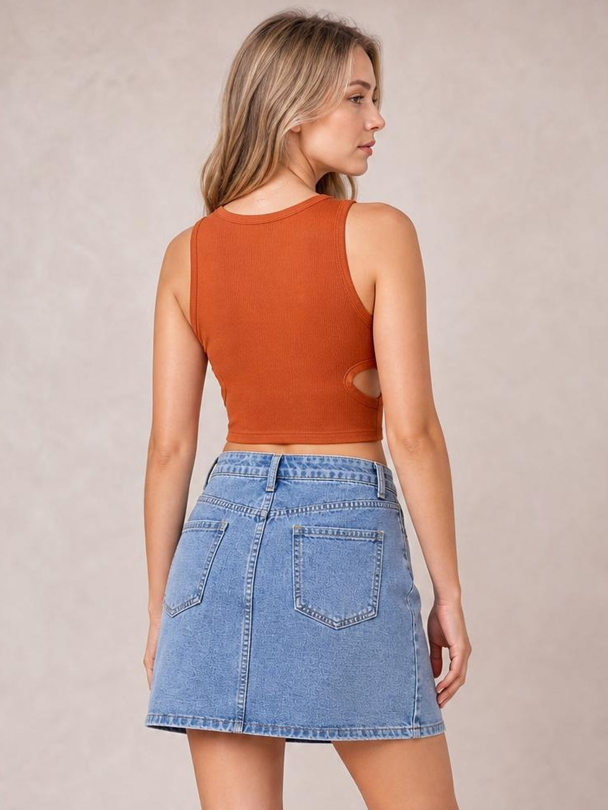 Model shown from behind wearing a light-blue denim mini skirt with a structured silhouette, belt loops, contrast stitching, back pockets, and an asymmetrical hem.