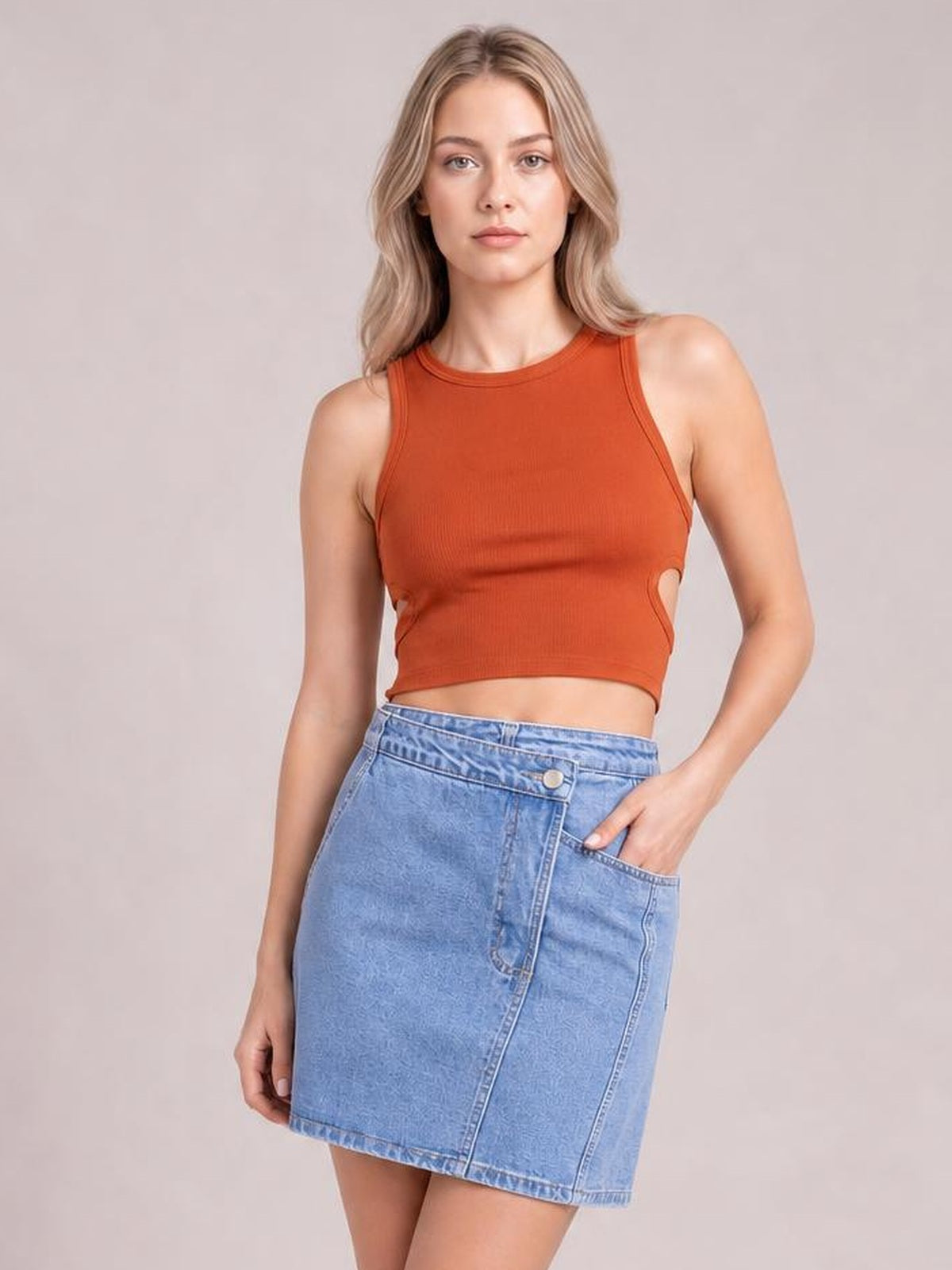 A model poses in a light blue asymmetrical denim mini skirt with button details, a wrap-style front panel, structured seams, and a straight silhouette.