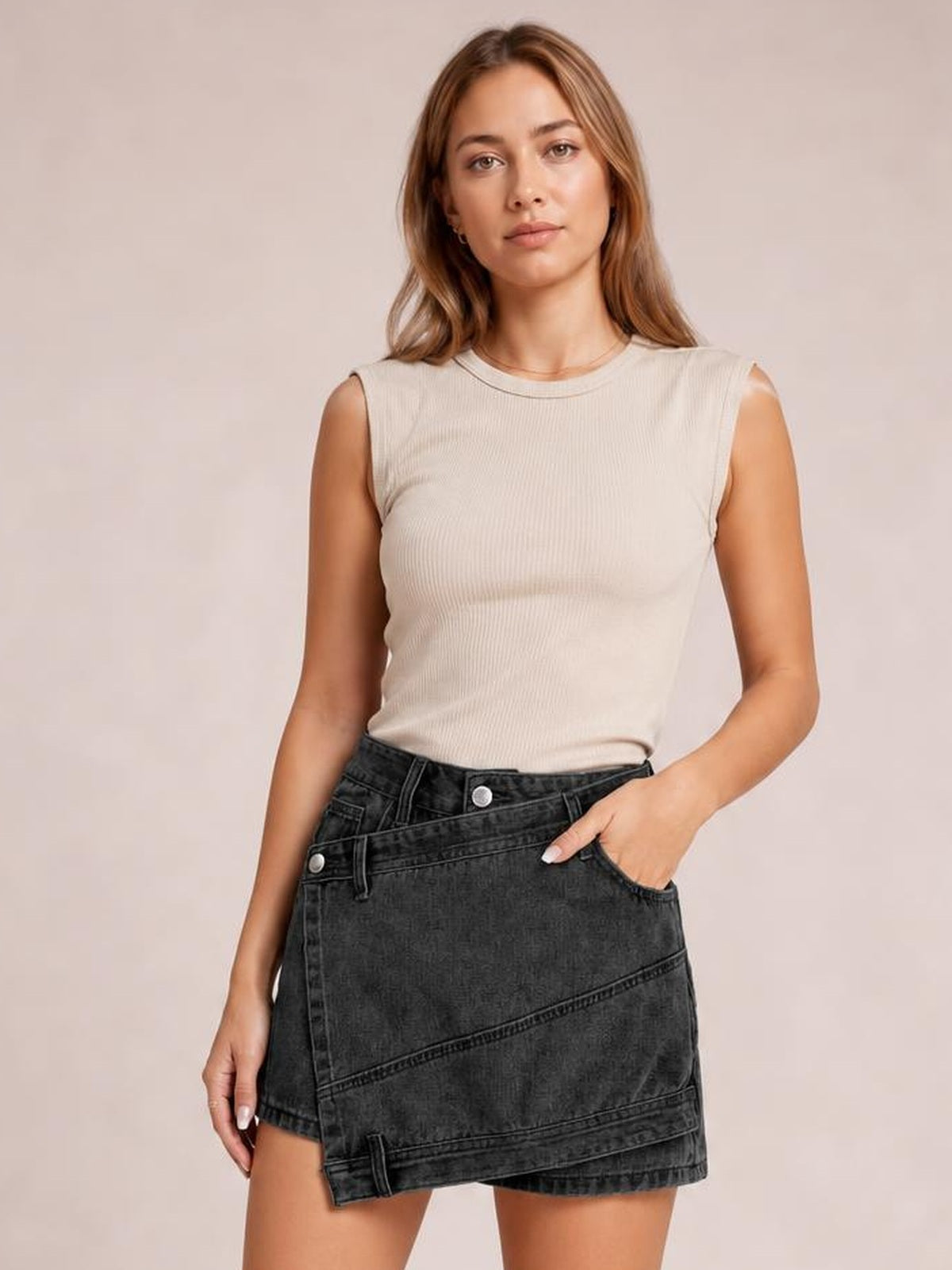 A model poses with one hand in a pocket, wearing a dark charcoal denim skort with an asymmetrical wrap-style front, diagonal seams, and a structured short silhouette.