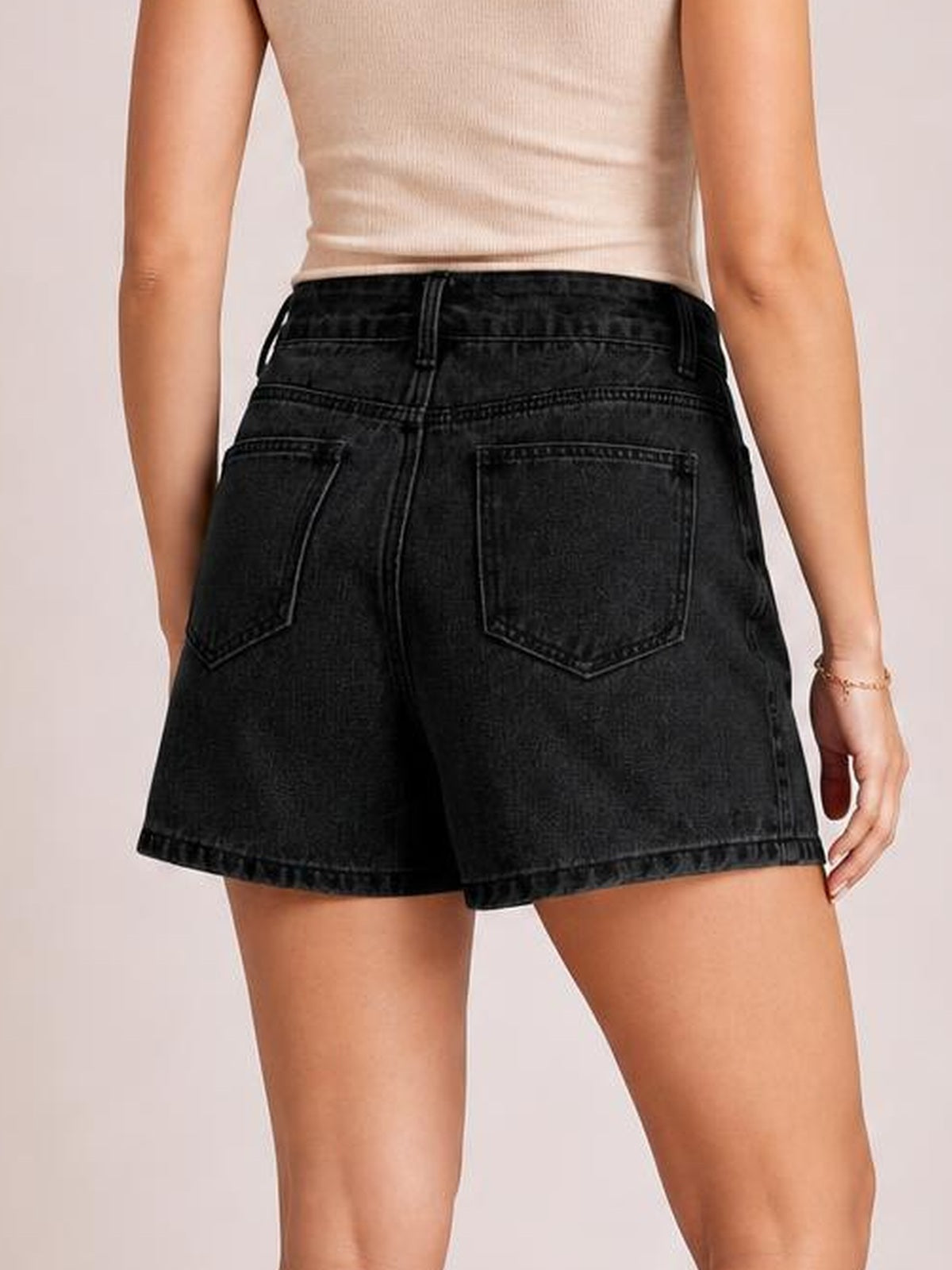 Model shown from behind wearing a black asymmetrical denim skort with a structured high waist, belt loops, stitched back pockets, and a relaxed short silhouette.