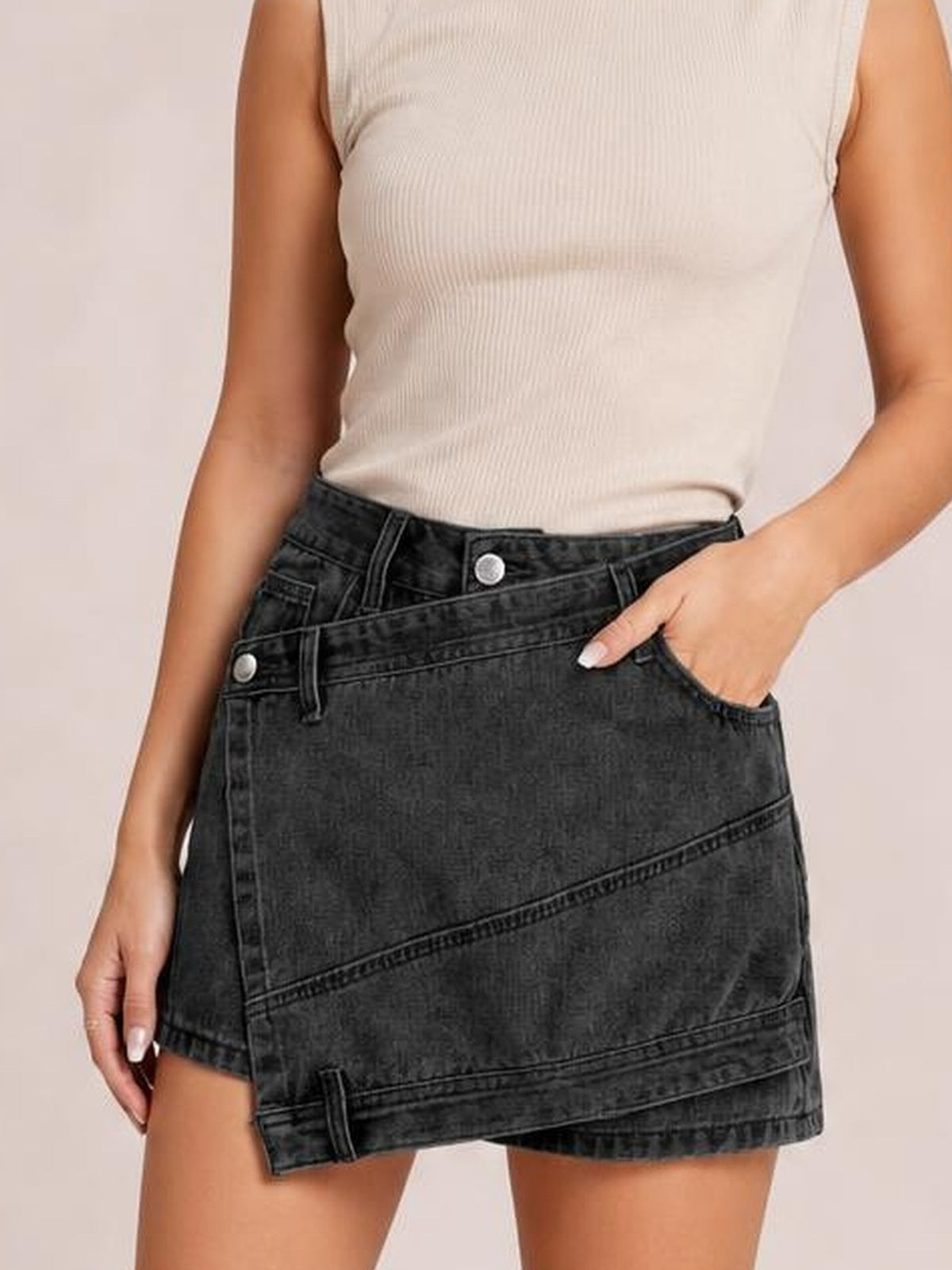 Model wears a structured dark denim skort with an asymmetrical overlapping front, diagonal seam detailing, belt loops, button closure, and pockets.