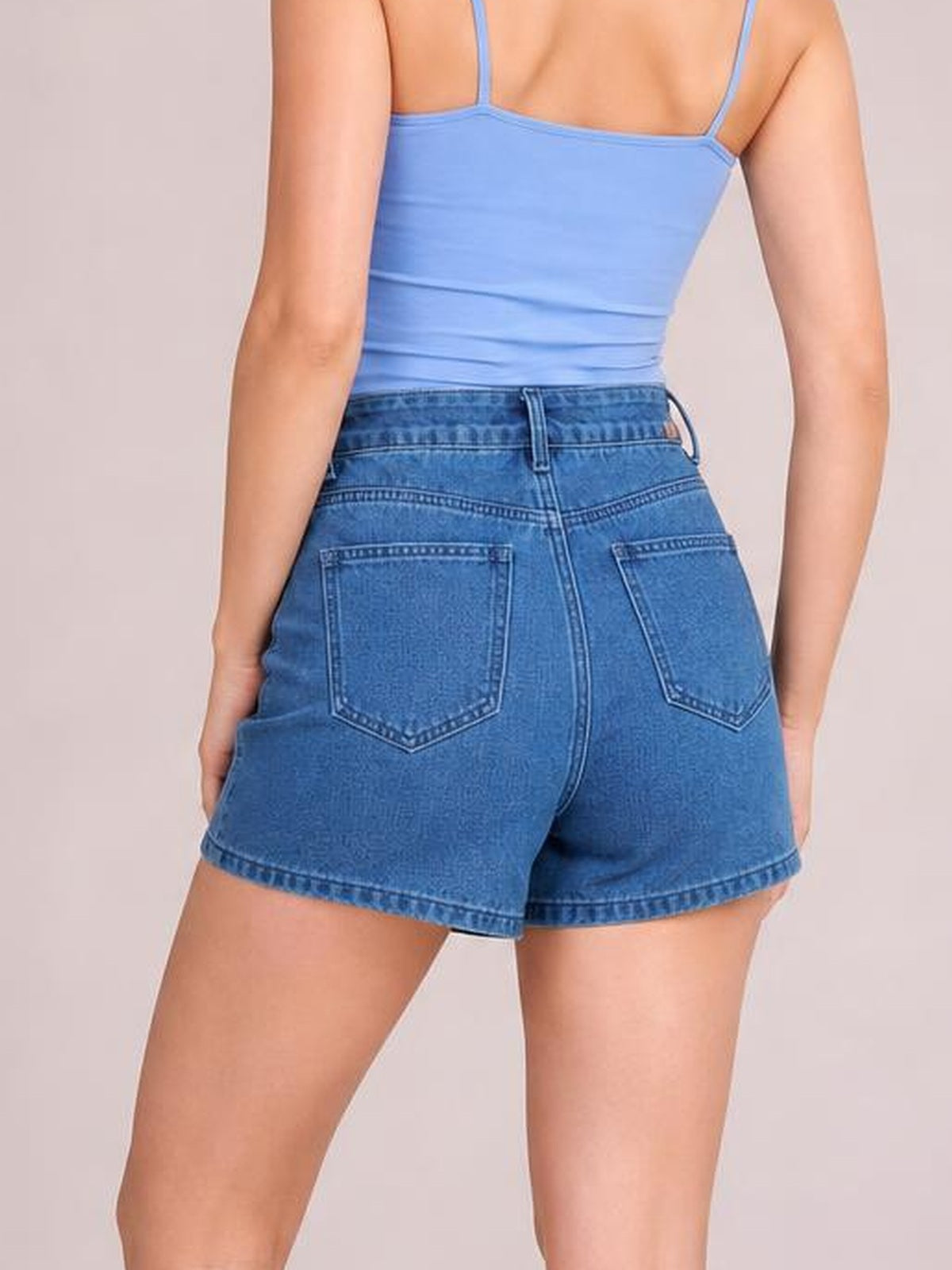 A model poses from behind wearing a blue asymmetrical denim skort with a structured waistband, belt loops, stitched back pockets, and a relaxed short silhouette.