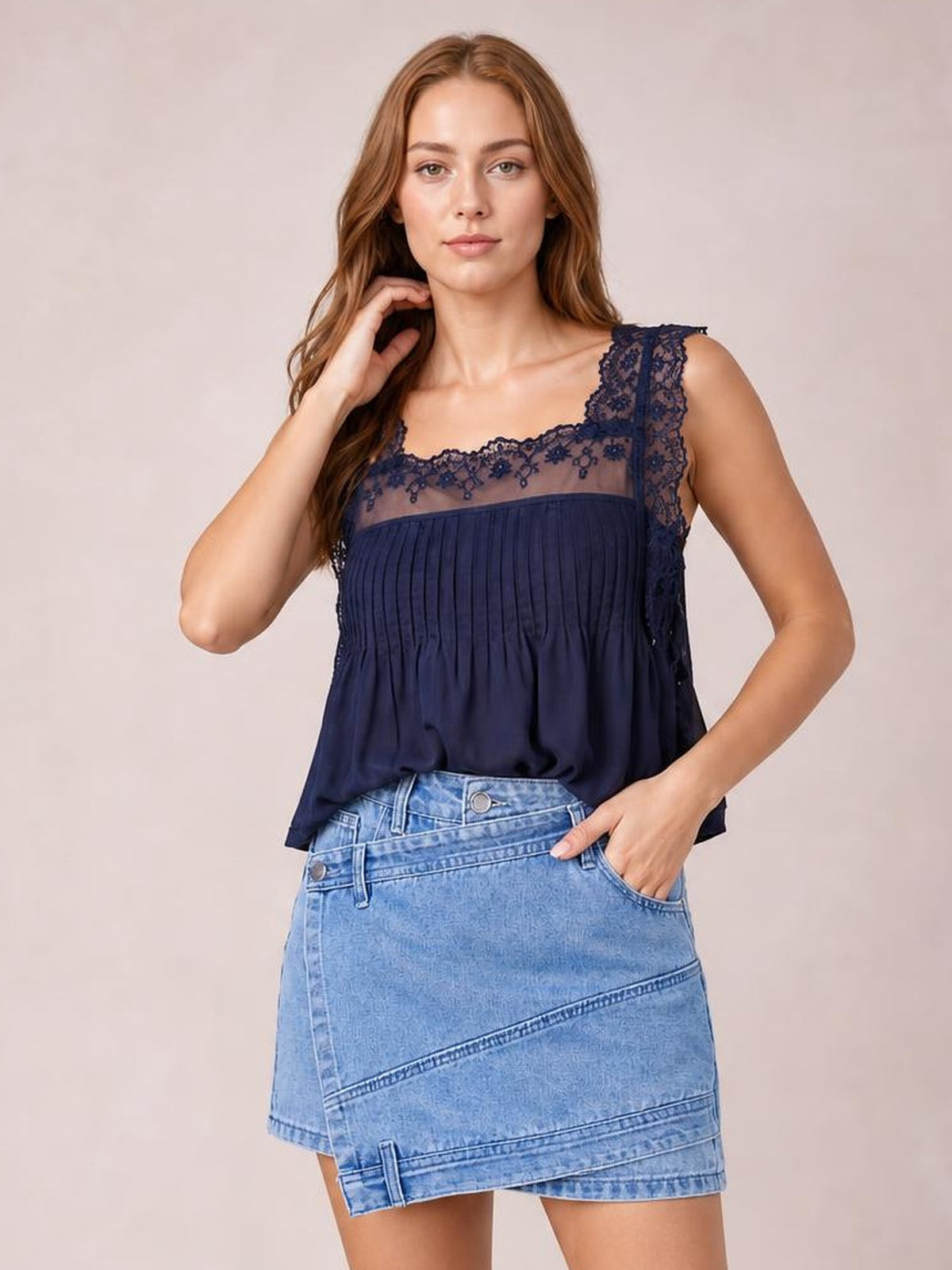 Model poses with one hand in a pocket, wearing a light-wash asymmetrical denim skort with a structured wrap front, diagonal seams, and layered hem.