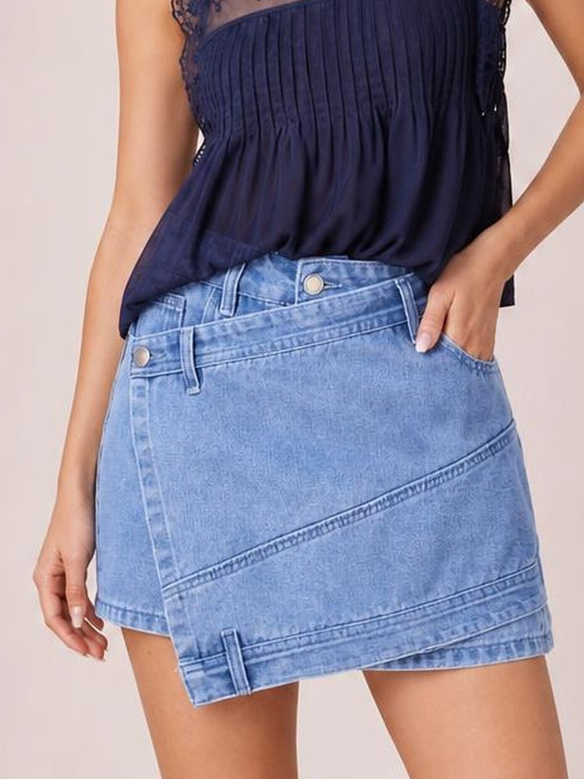 A model wears a light-blue asymmetrical denim skort with front pockets, a structured waistband, diagonal panel seams, and shorts visible beneath the skirt hem.