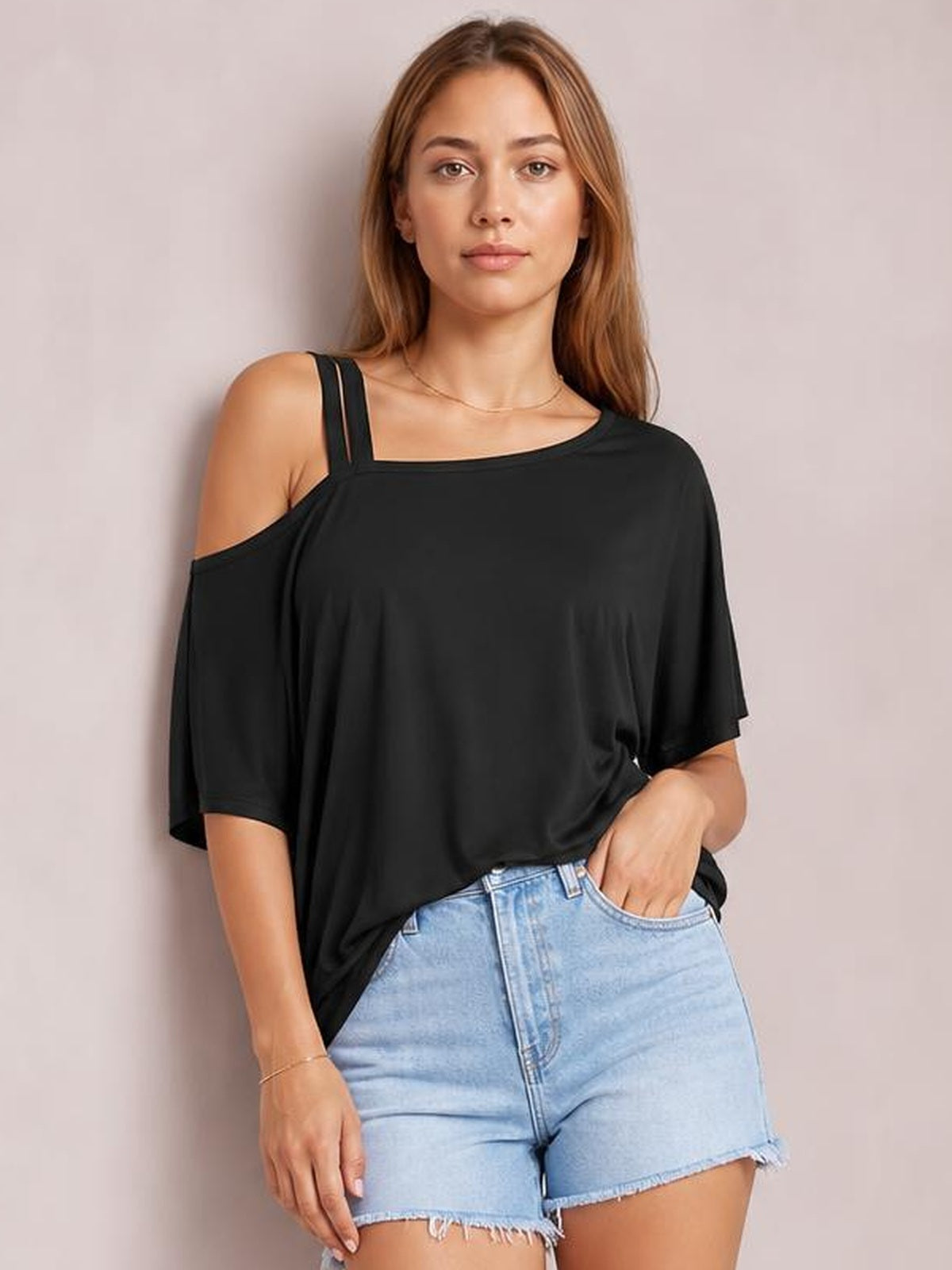 Model poses in a loose black asymmetrical-neck half-sleeve T-shirt with an exposed shoulder and draped silhouette, one hand in her shorts pocket.