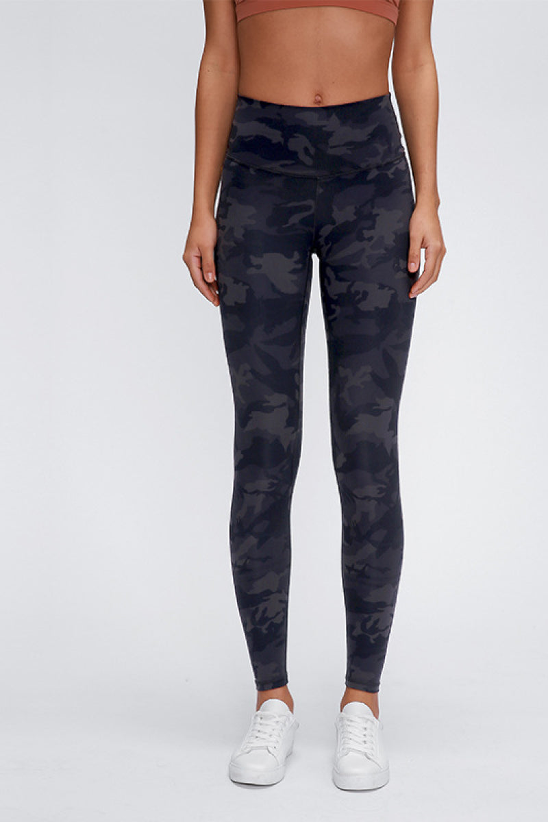 Model stands front-facing in fitted dark camouflage sports leggings with a wide seamless band waist and full-length tapered silhouette.