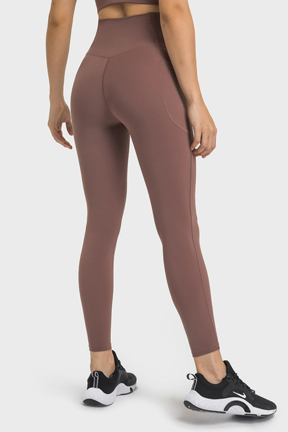 A model stands in brown V-waist yoga leggings with side pockets, a smooth high-rise fit, curved seams, and a close ankle-length silhouette.