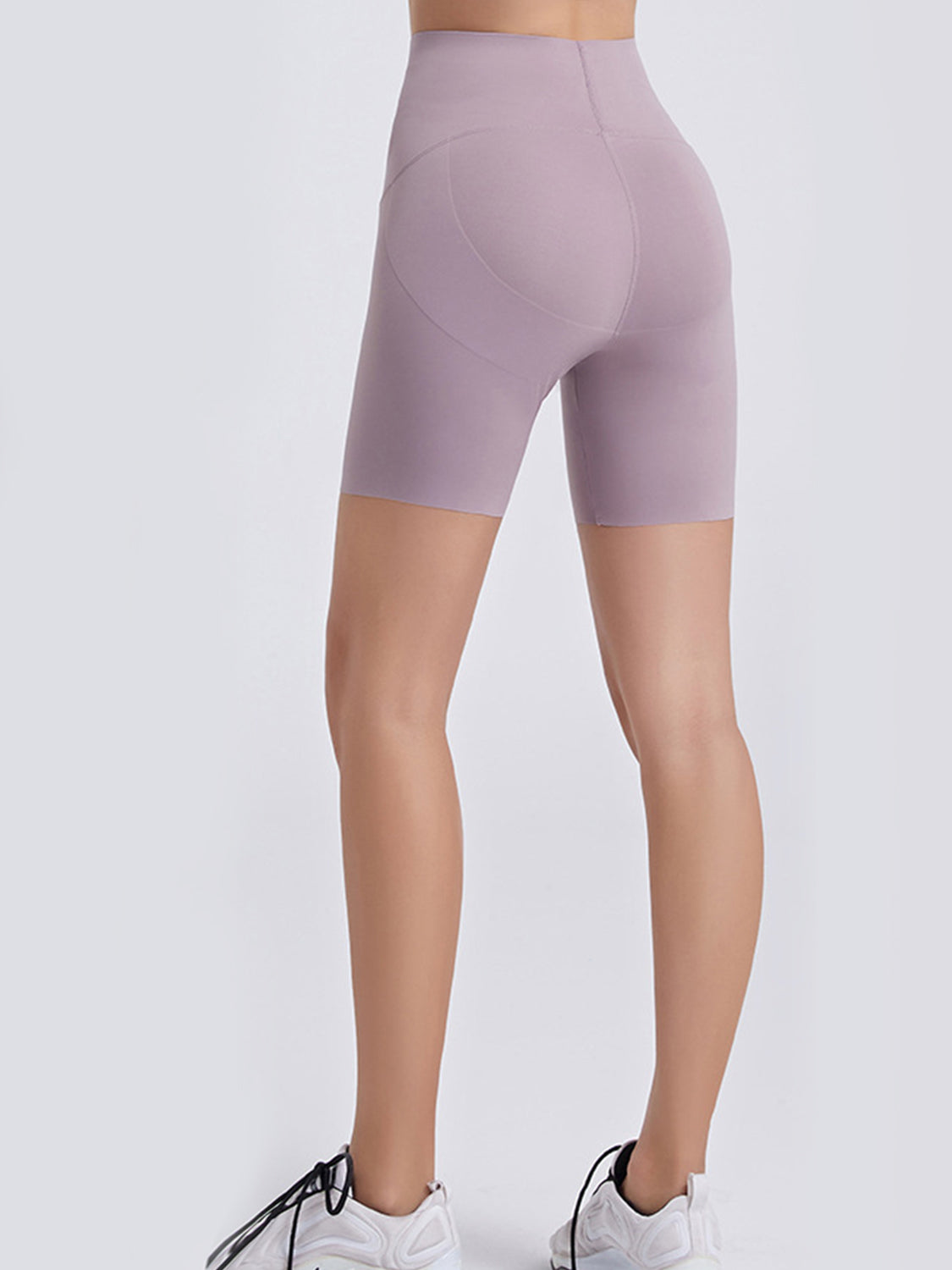 Back view of a model standing in lavender wide-waistband sports shorts with a high-rise fit, stretch silhouette, mid-thigh length, and curved panel seams.