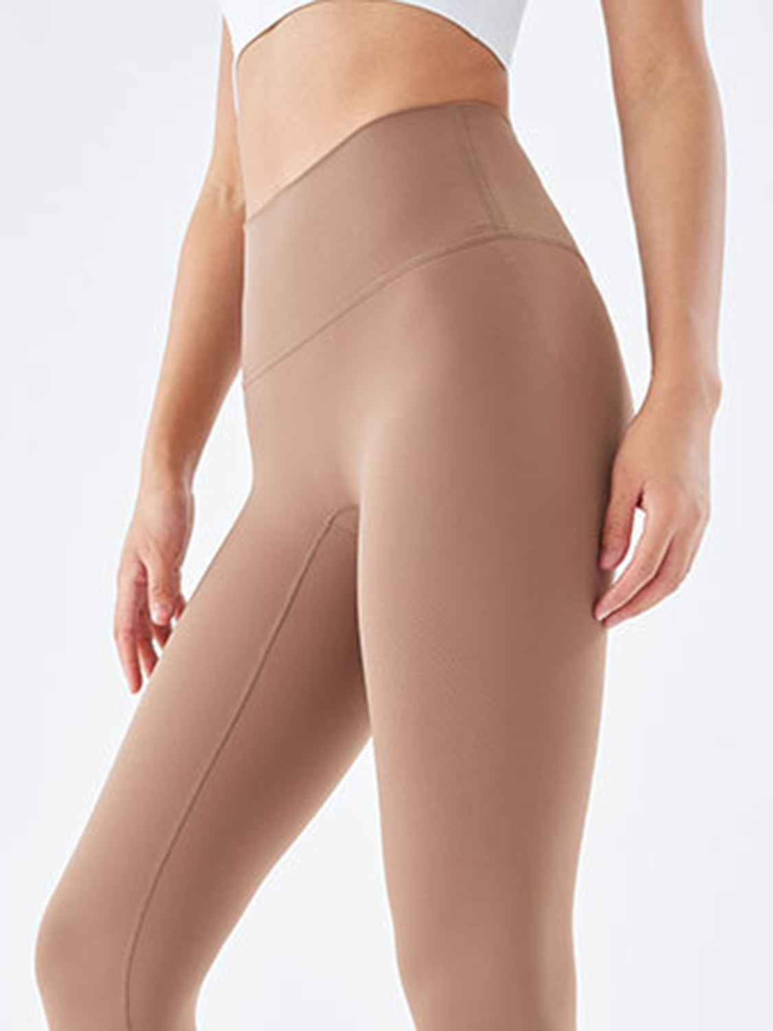 Model wearing tan wide waistband sports leggings with a high-rise, close-fitting silhouette, visible leg seams, and a relaxed stance with hands resting by her thighs.