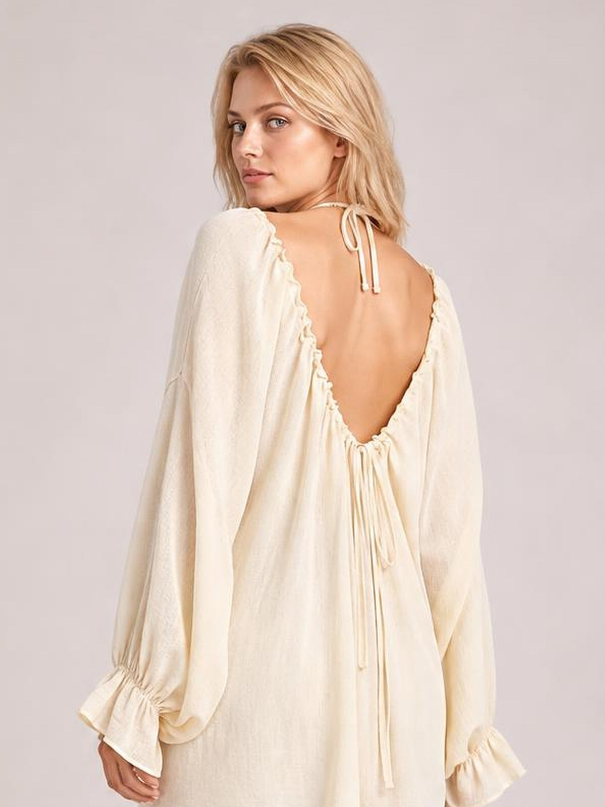Model looks over her shoulder in an ivory backless cover-up with a deep drawstring back, loose silhouette, and long flounce sleeves.
