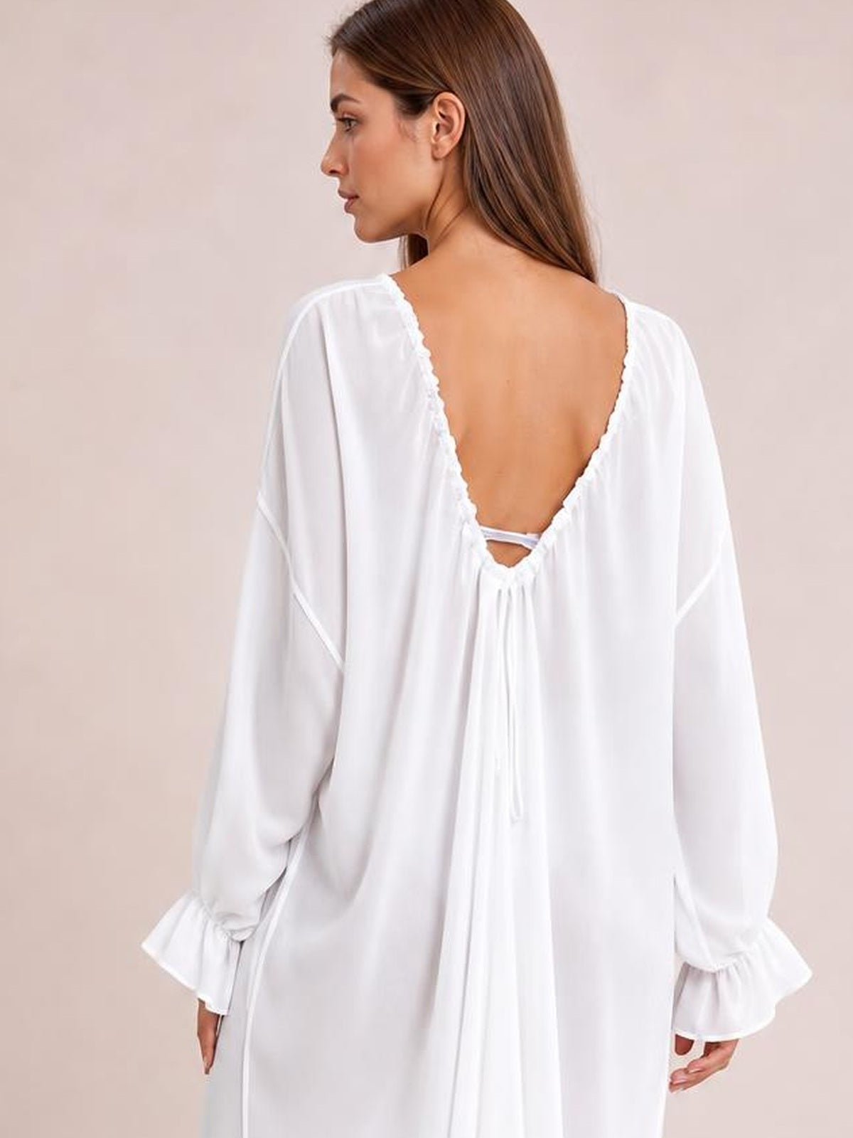 Model shown from the back in a white backless cover-up with an adjustable drawstring, flowing silhouette, and long flounce sleeves.