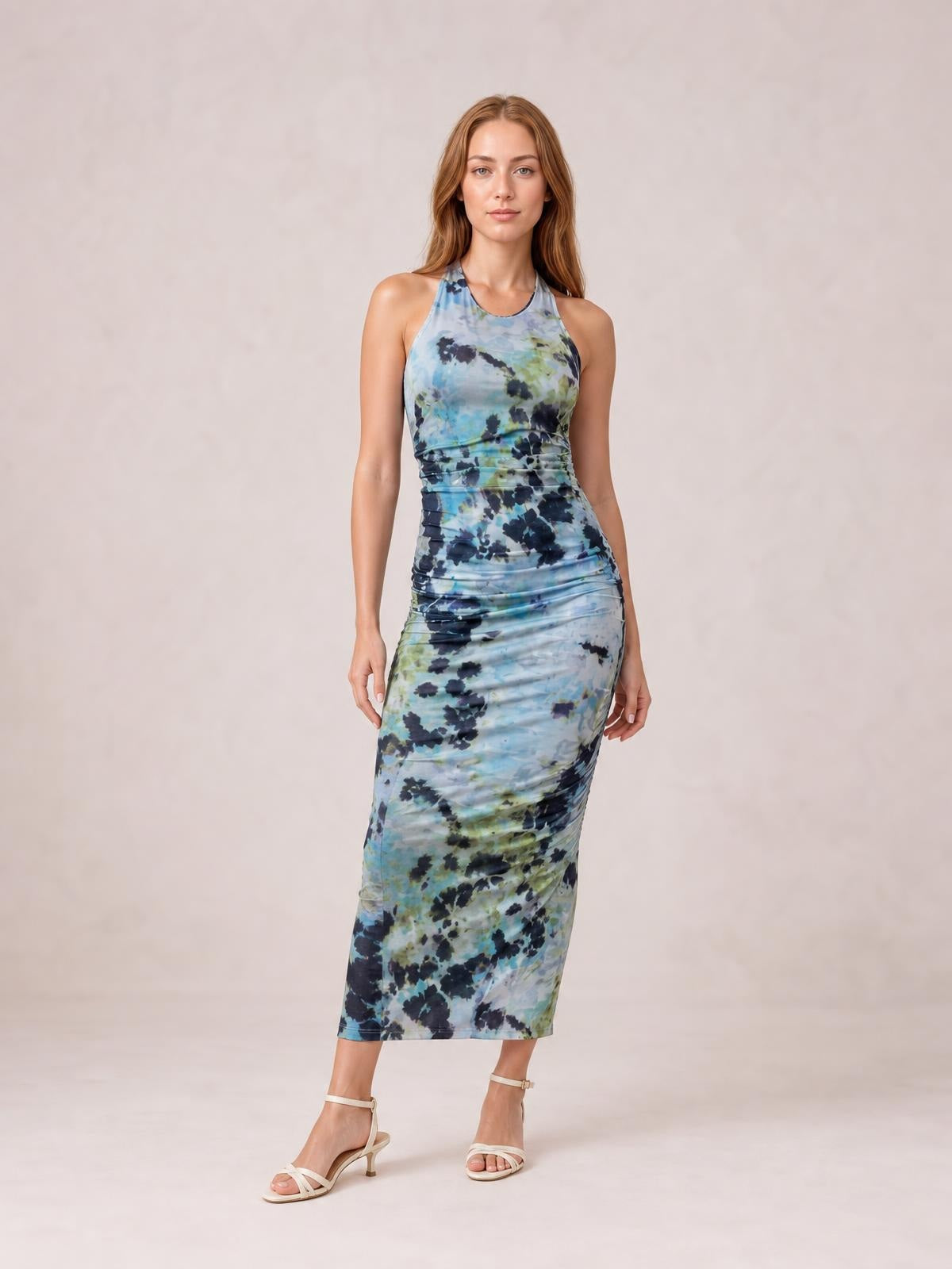 Model poses in a blue, green, and black printed halter neck dress with a fitted ruched silhouette and midi-length hem.