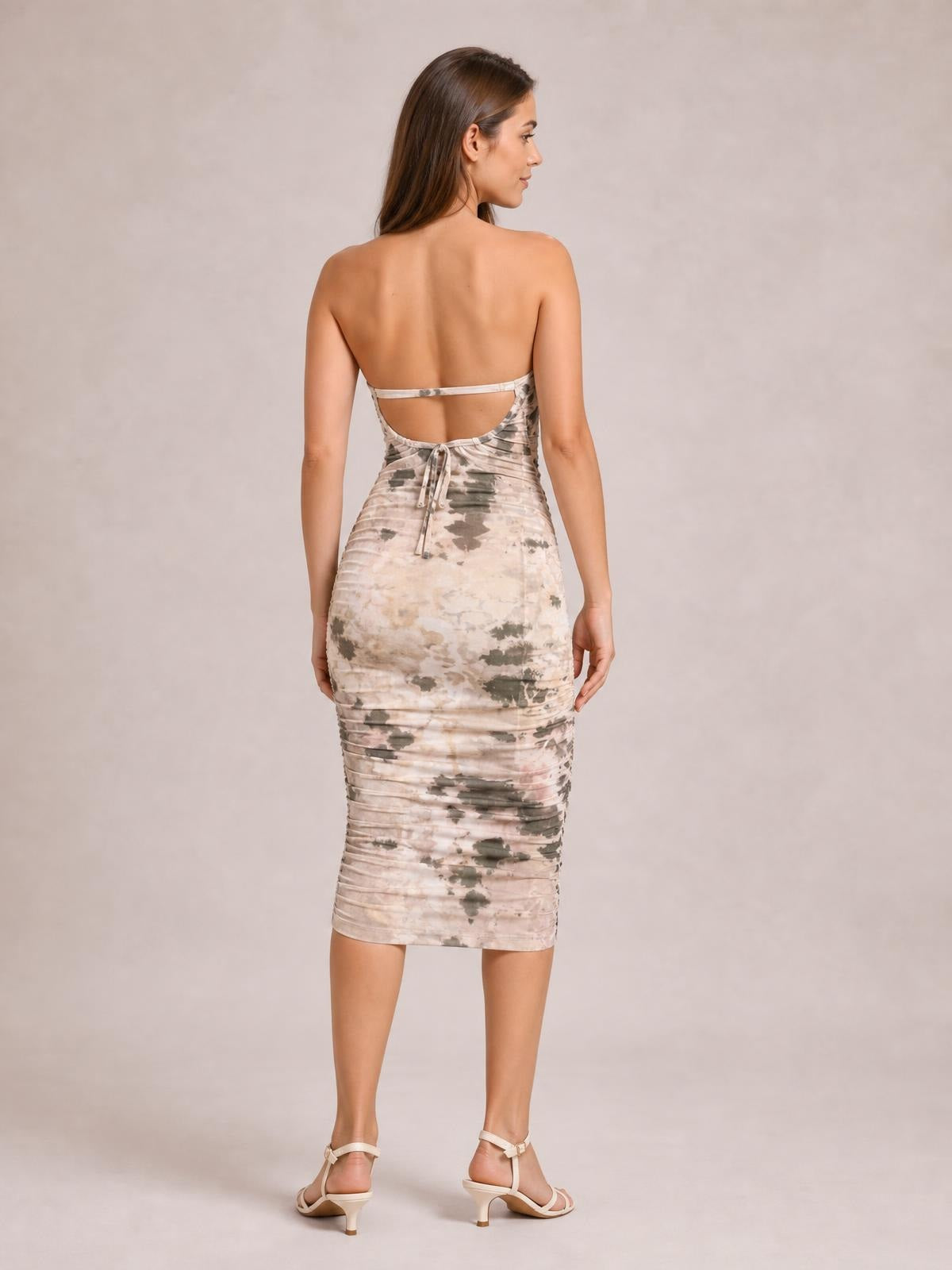 Model shown from behind in a fitted printed halter neck midi dress with a pale multicolor pattern, open back, back tie, and ruched silhouette.