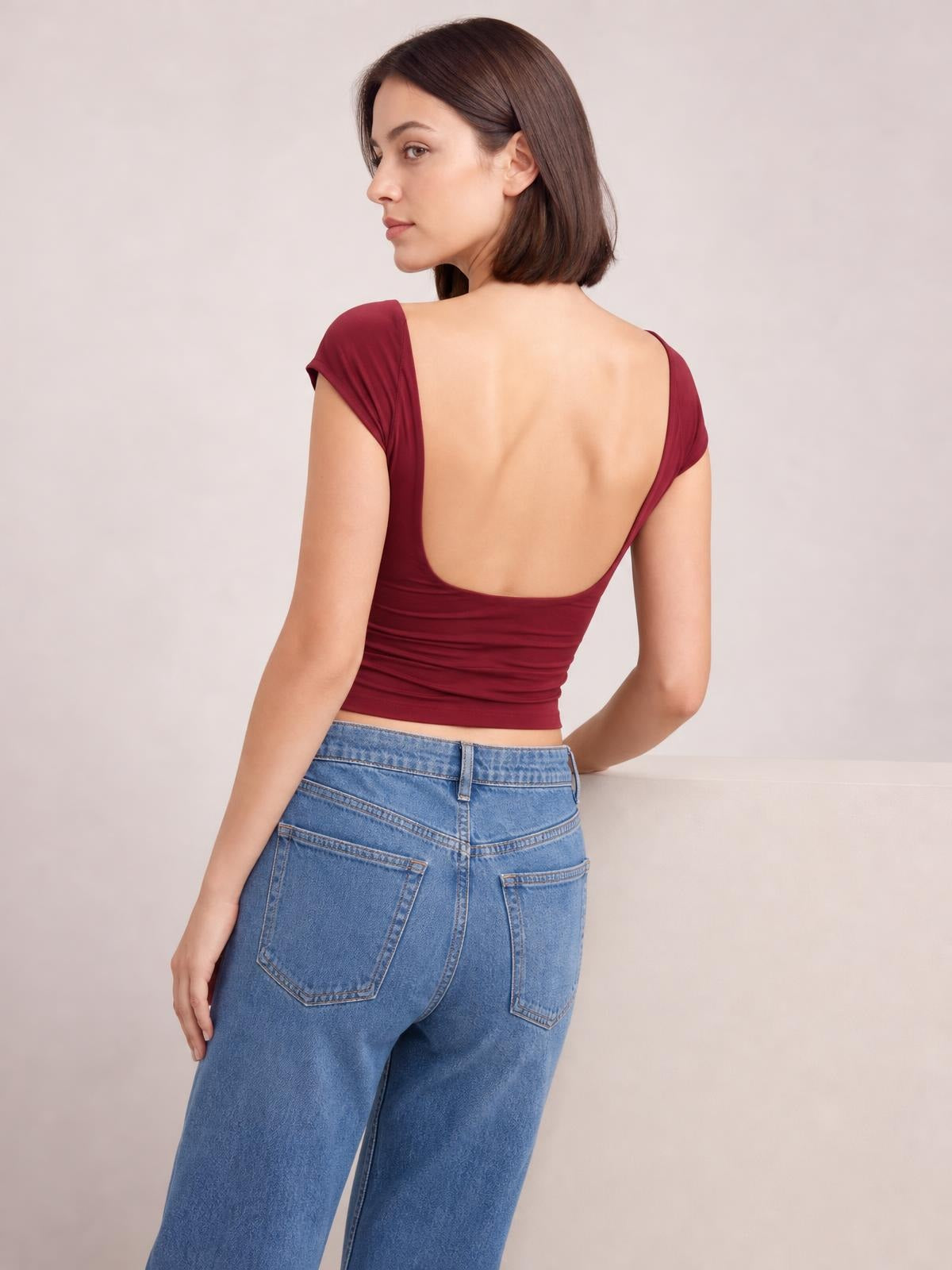 Model in a burgundy solid cropped blouse poses in profile, showing its short sleeves, fitted silhouette, round neckline, and deep backless cutout.