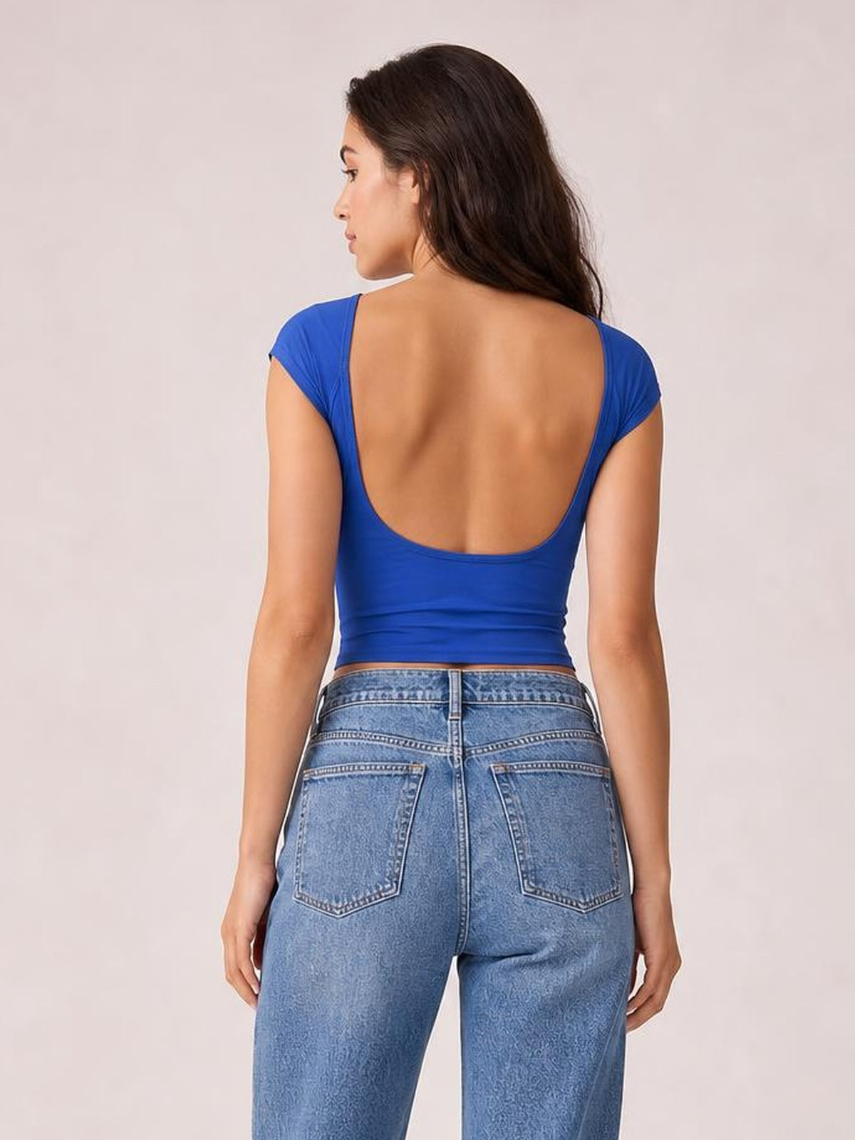 Model shown from the back wearing a solid blue cropped blouse with short sleeves, a rounded neckline, and a deep curved back cutout.