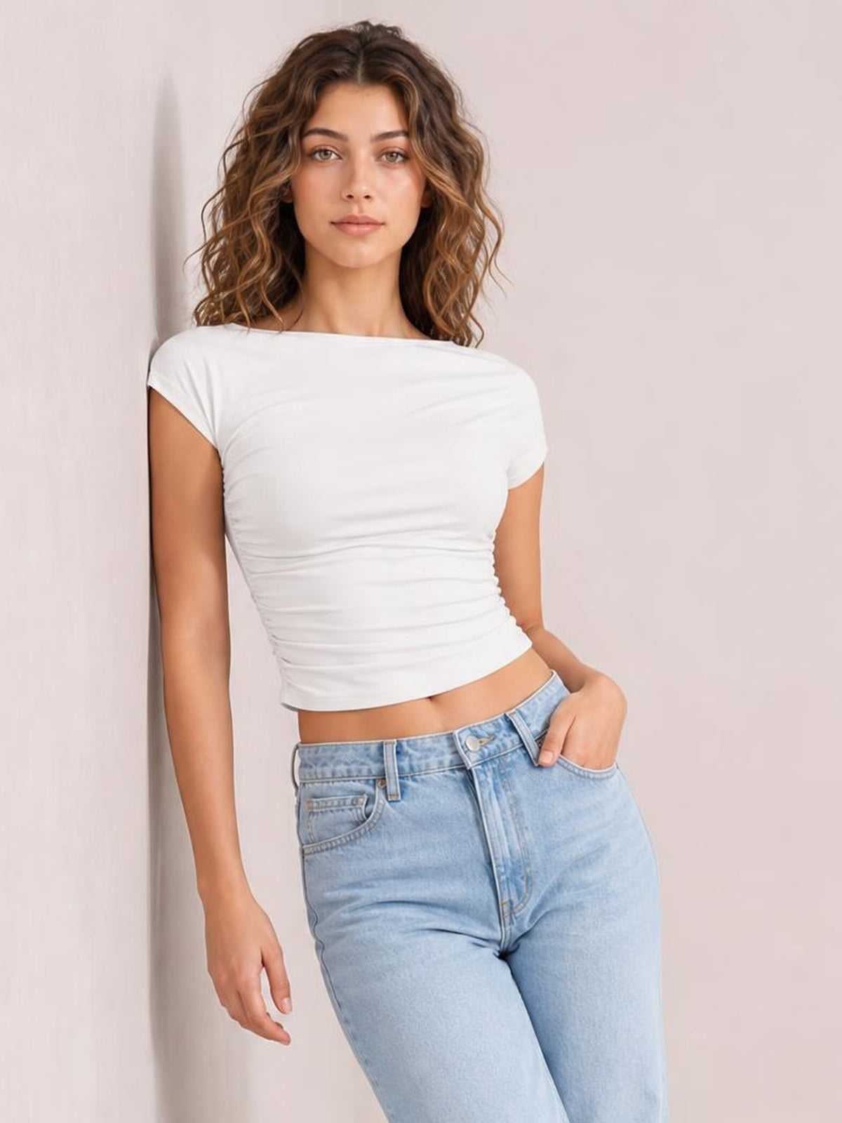 Model wears a solid white cropped blouse with a round neckline, short regular sleeves, fitted ruched sides, and a casual silhouette while posing with one hand in her jeans pocket.
