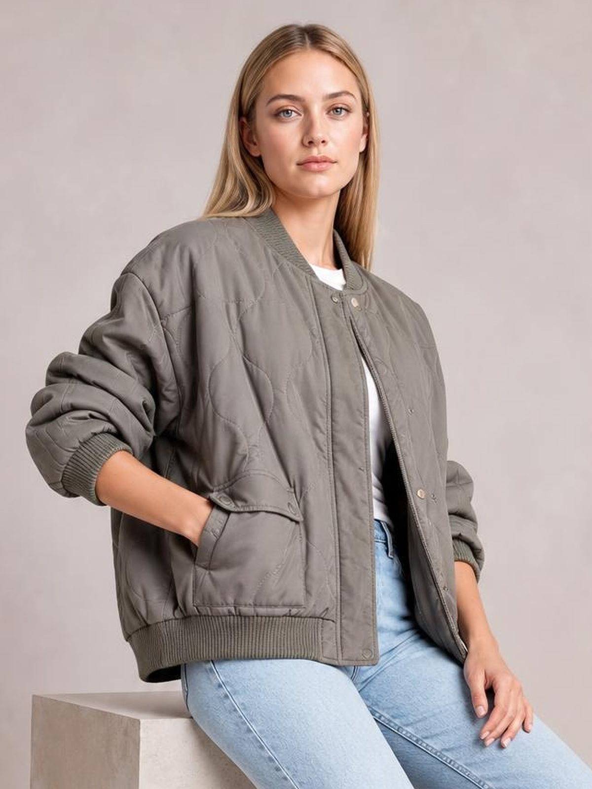 A seated model wears a gray snap-down long sleeve jacket with a ribbed baseball collar, cuffs and hem, relaxed bomber silhouette, and front pockets.