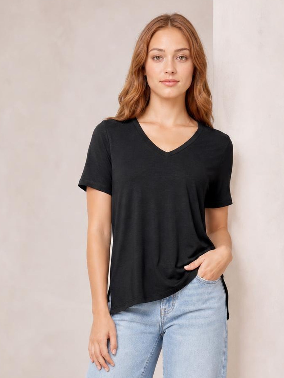Model wearing a black bamboo V-neck T-shirt with short sleeves, a relaxed silhouette, and a high-low hem, posing with one hand in her jeans pocket.