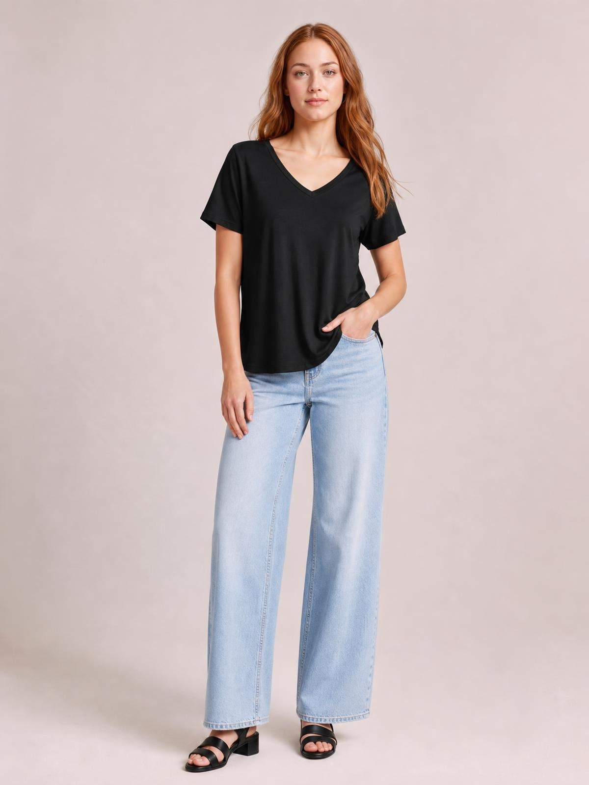 Model wears a black bamboo V-neck T-shirt with short sleeves, a relaxed silhouette, and a curved high-low hem while posing with one hand in her jeans pocket.