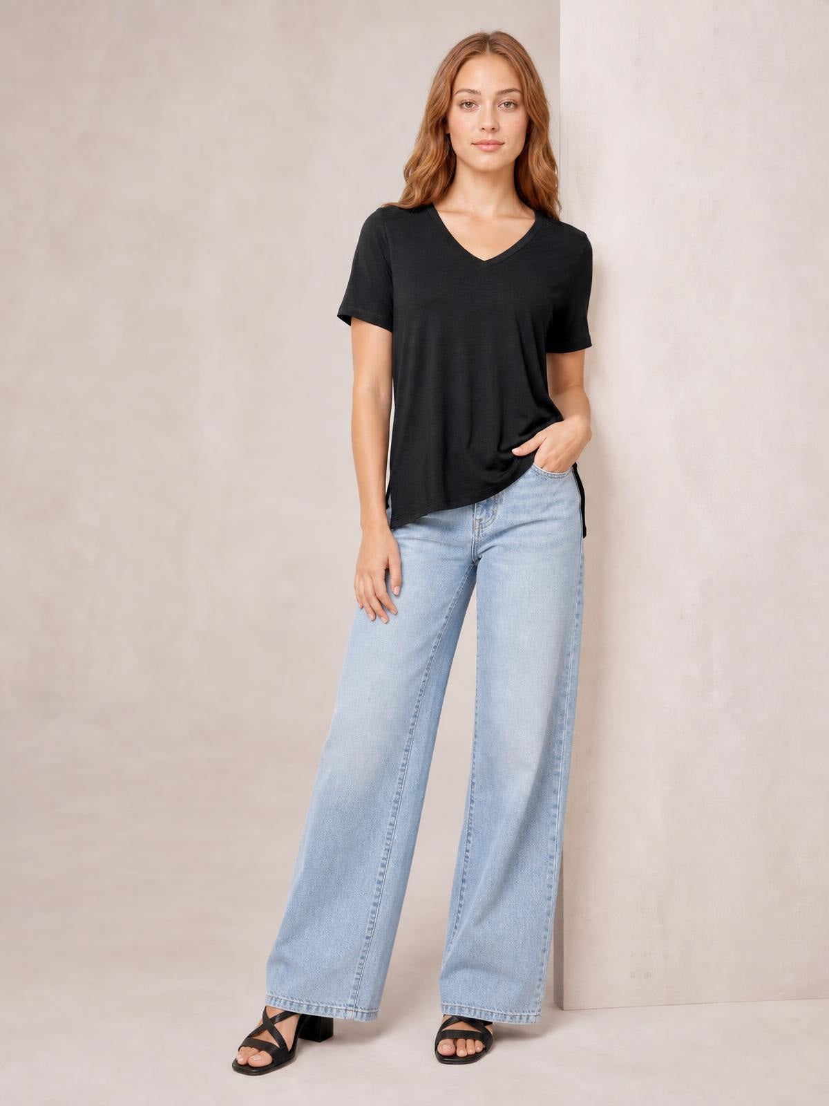 Model wears a black bamboo V-neck T-shirt with short sleeves and a relaxed high-low hem, posed with one hand in her jeans pocket.