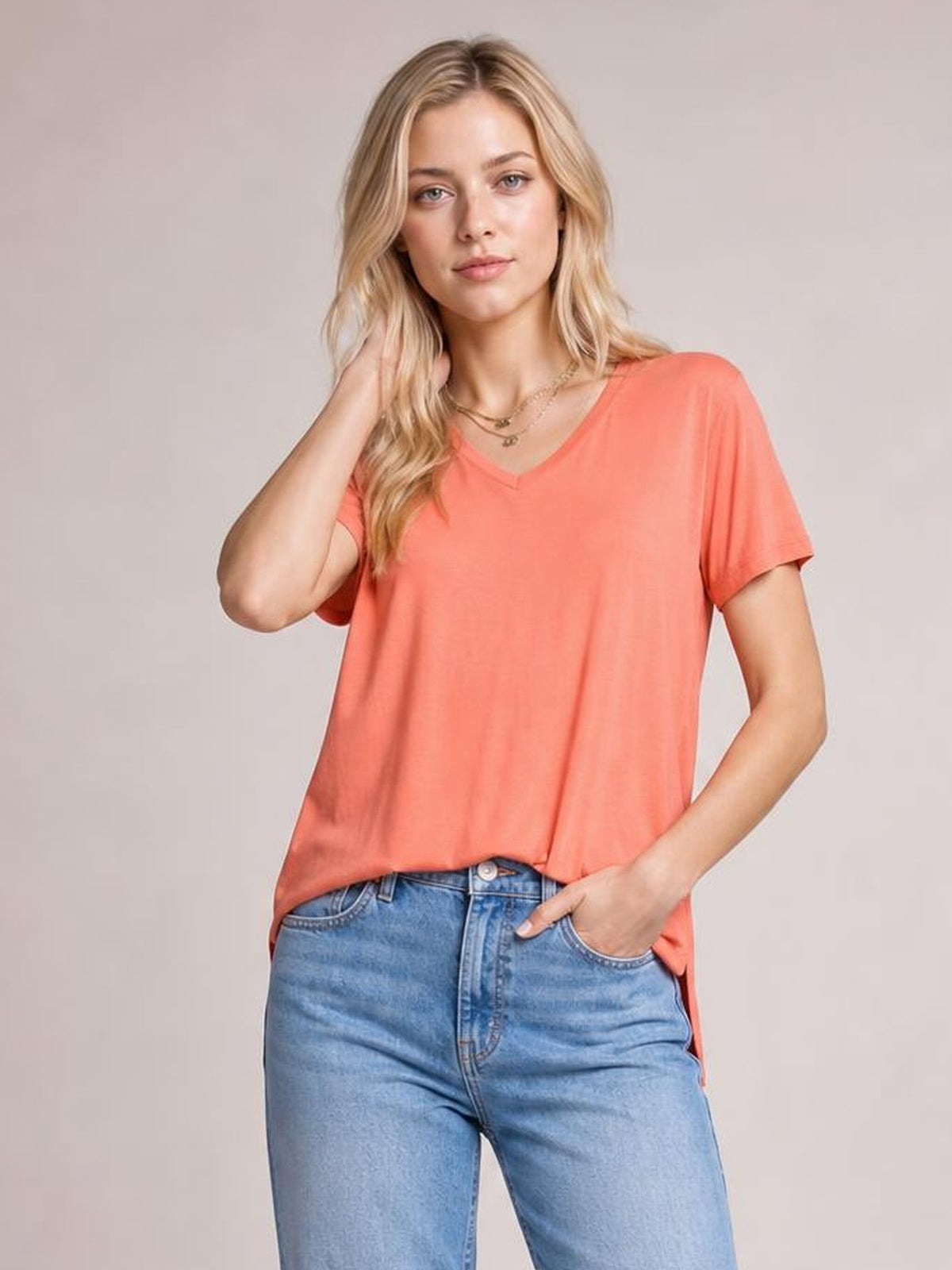 Model poses in a coral bamboo V-neck T-shirt with short sleeves, a relaxed silhouette, and a high-low hem, touching her hair with one hand and resting the other in her jeans pocket.