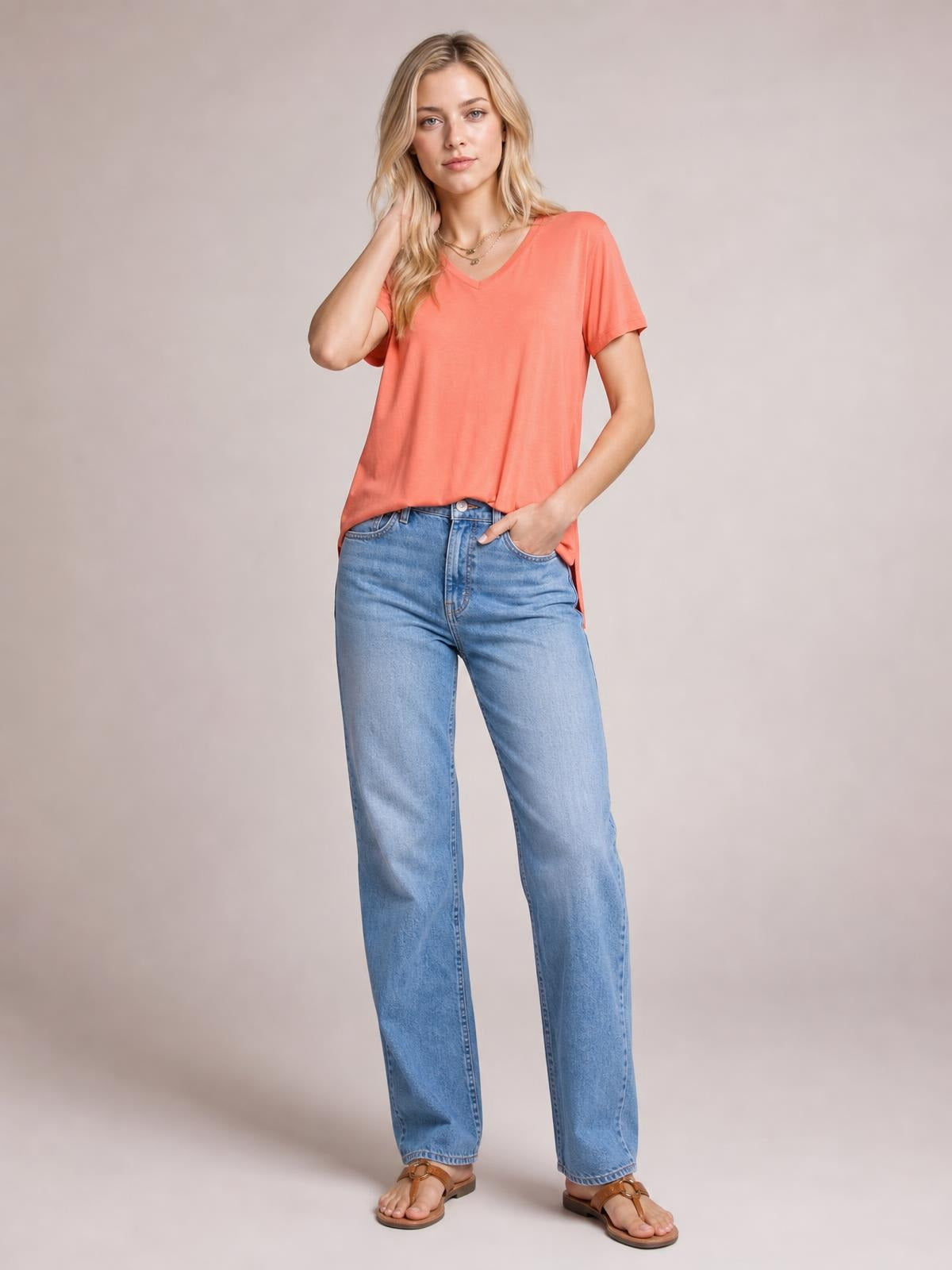 Model wears a coral bamboo V-neck T-shirt with short sleeves, relaxed silhouette, and high-low hem, posed with one hand in her jeans pocket.